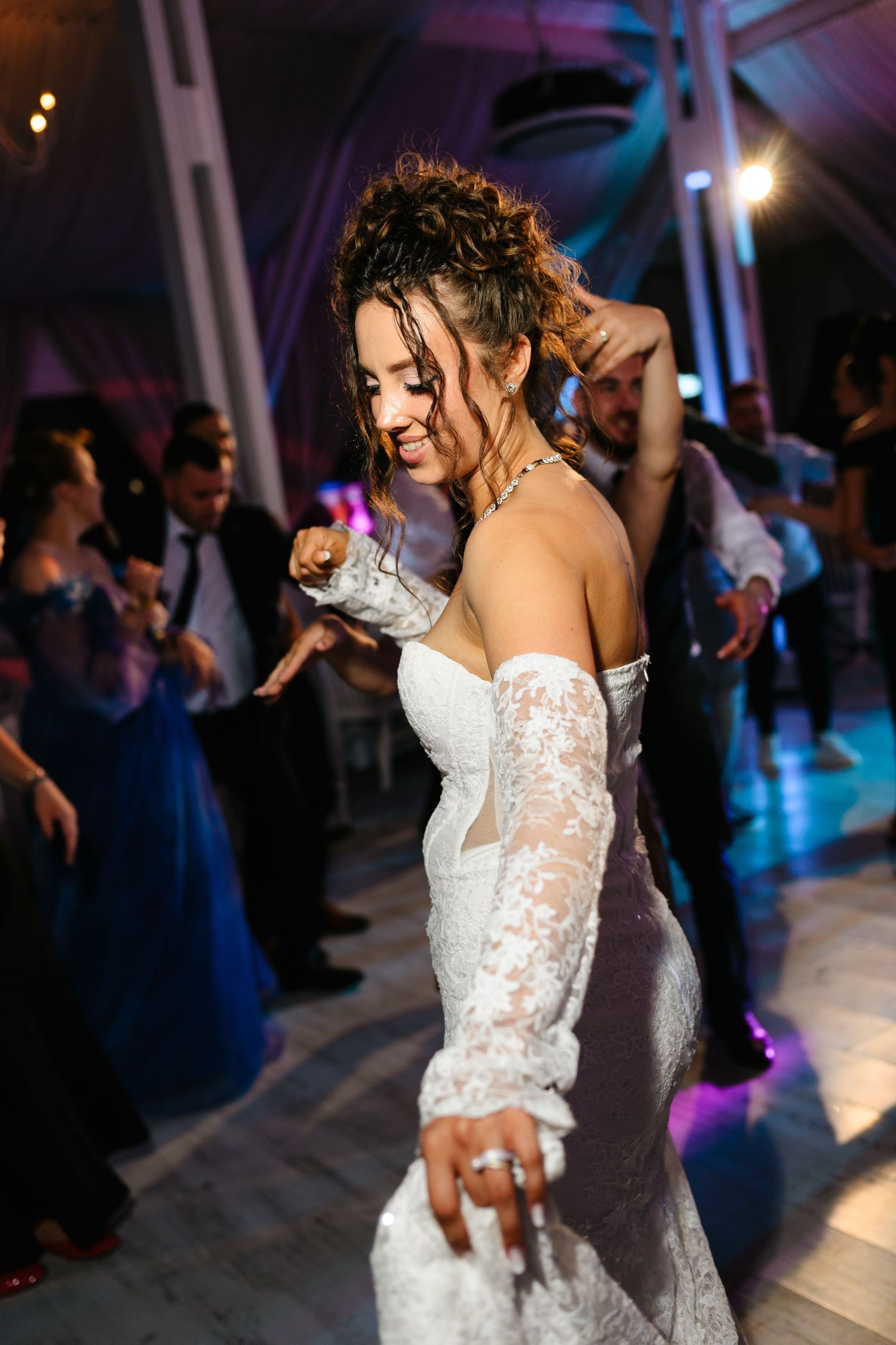 Bride in lace wedding gown dancing joyfully at wedding reception with guests celebrating in background.