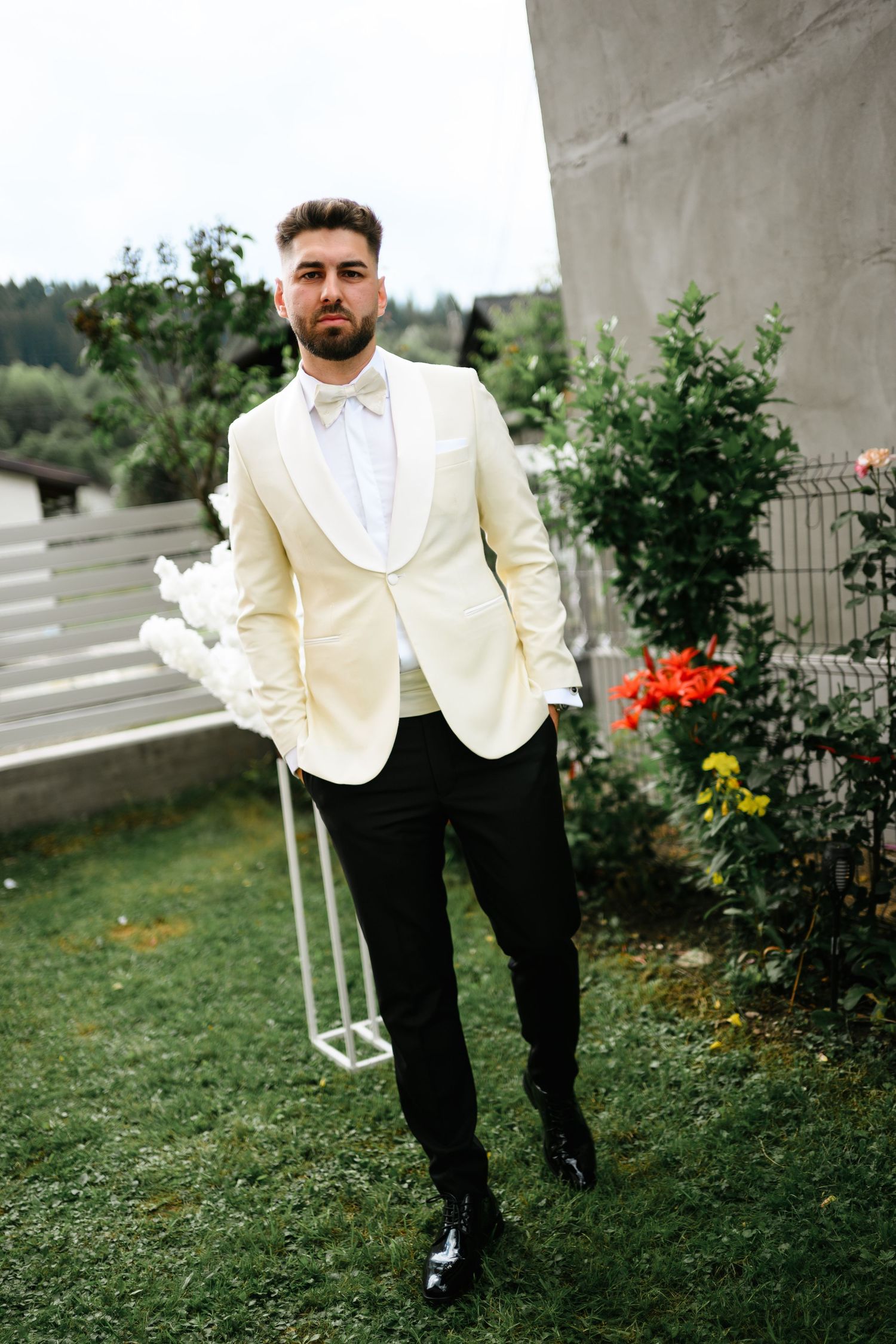 Groom in cream tuxedo jacket, black pants, and bow tie poses outdoors near flowers and greenery.