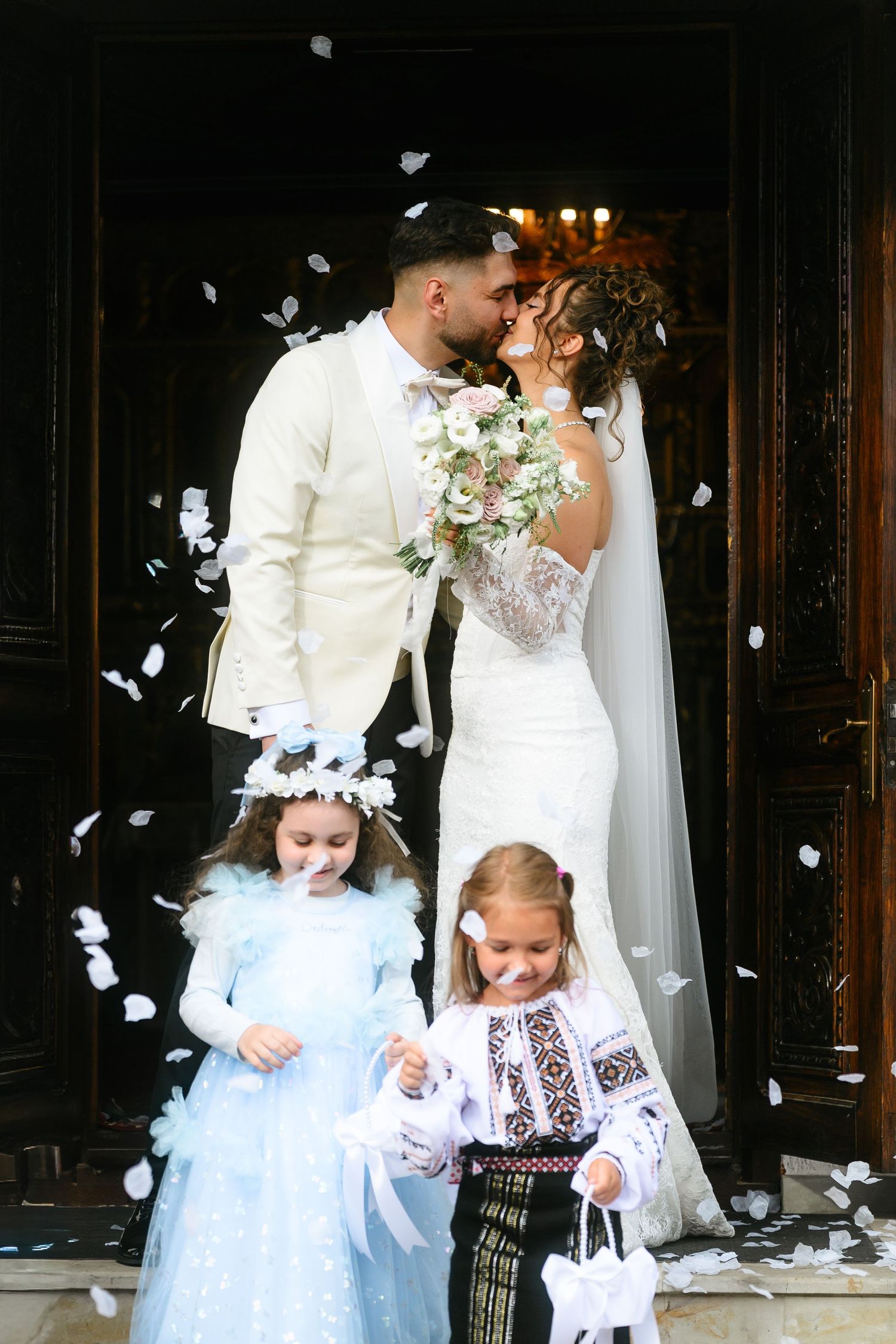 Bride and groom kissing at church doors with flower girls tossing white petals at a beautiful outdoor wedding ceremony.