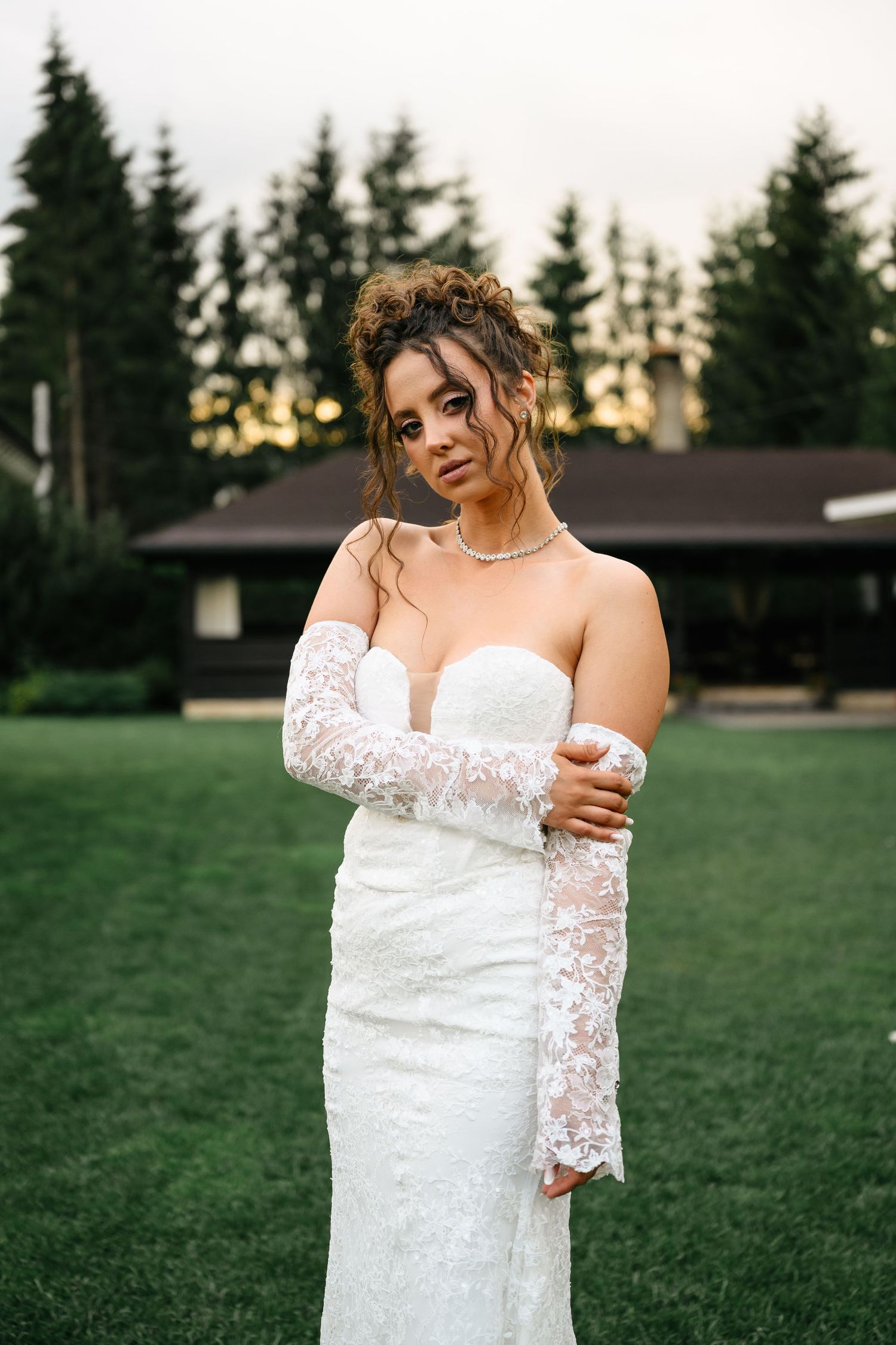 Bride in white lace off-shoulder gown with long sleeves poses on green lawn before evergreen trees at sunset.