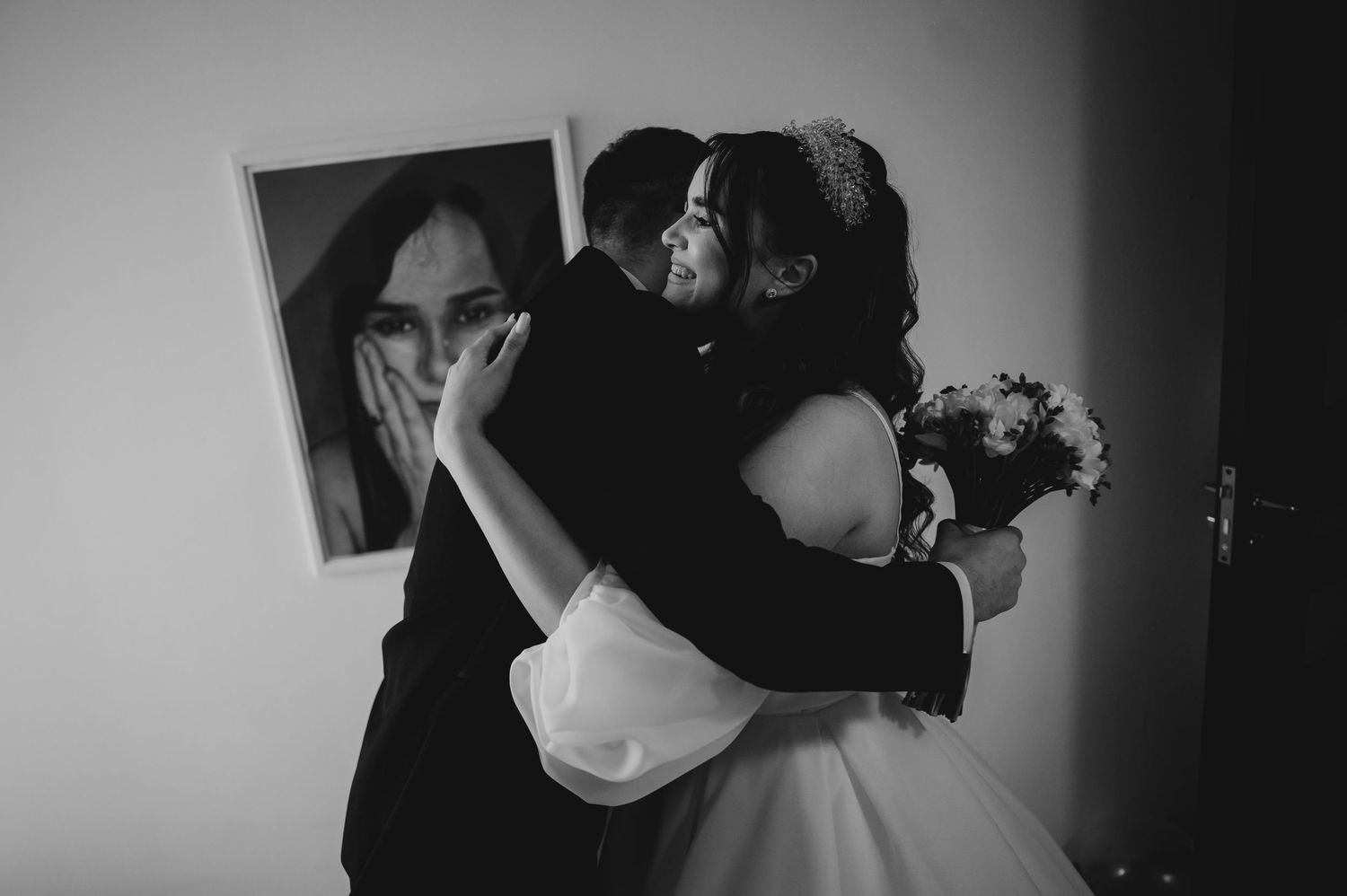 Black and white wedding photo of bride and groom sharing an emotional embrace, with a portrait hanging on the wall behind them.