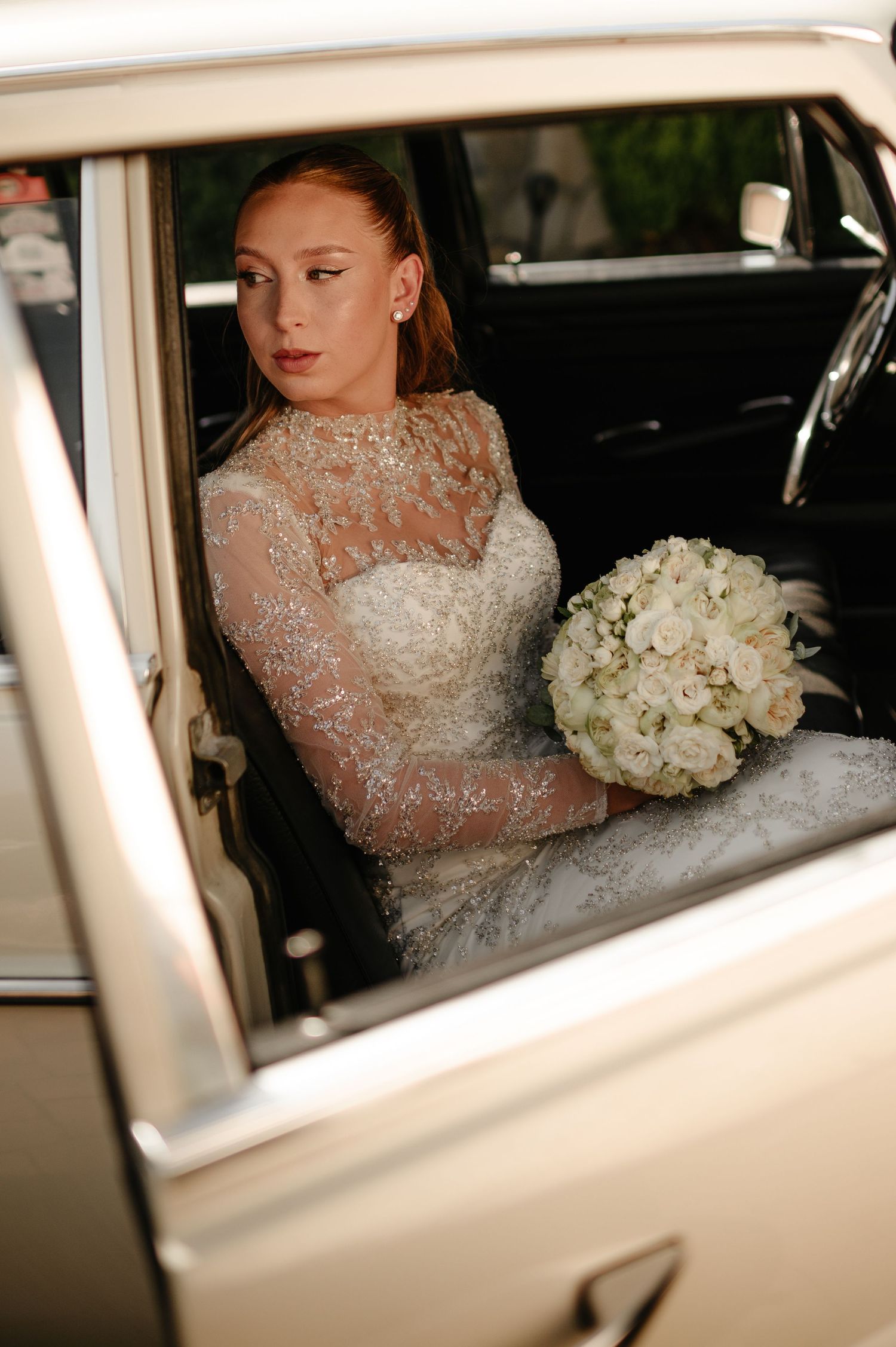 Elegant bride in beaded lace gown holding white bouquet seated in vintage classic car.