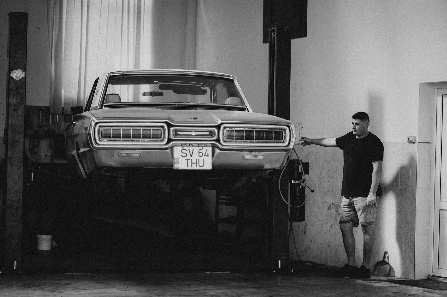 Black and white photo of a man standing beside a classic muscle car elevated on a garage lift.