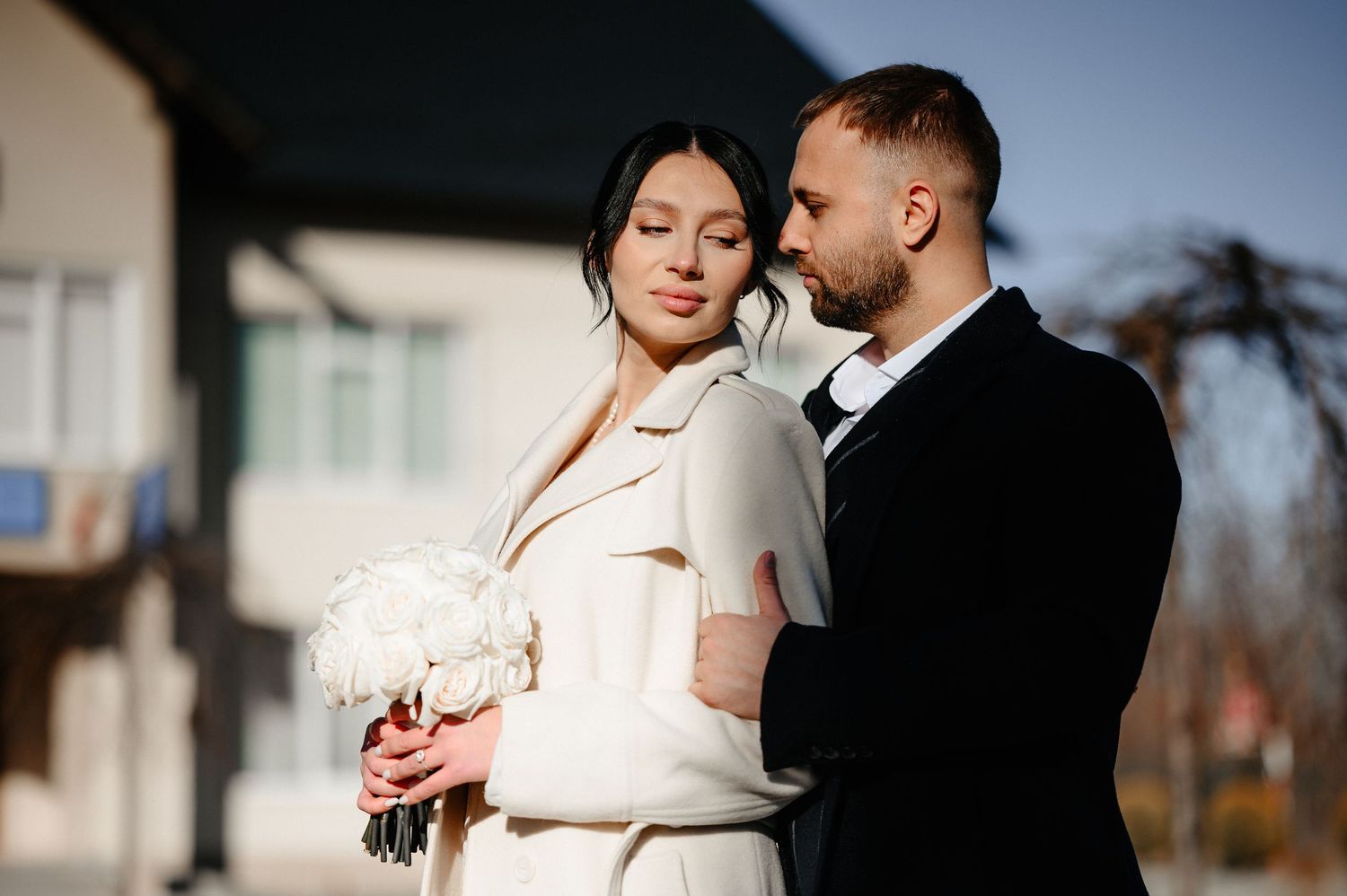Elegant bride and groom posing outdoors on sunny wedding day, bride holding white bouquet wearing cream coat.