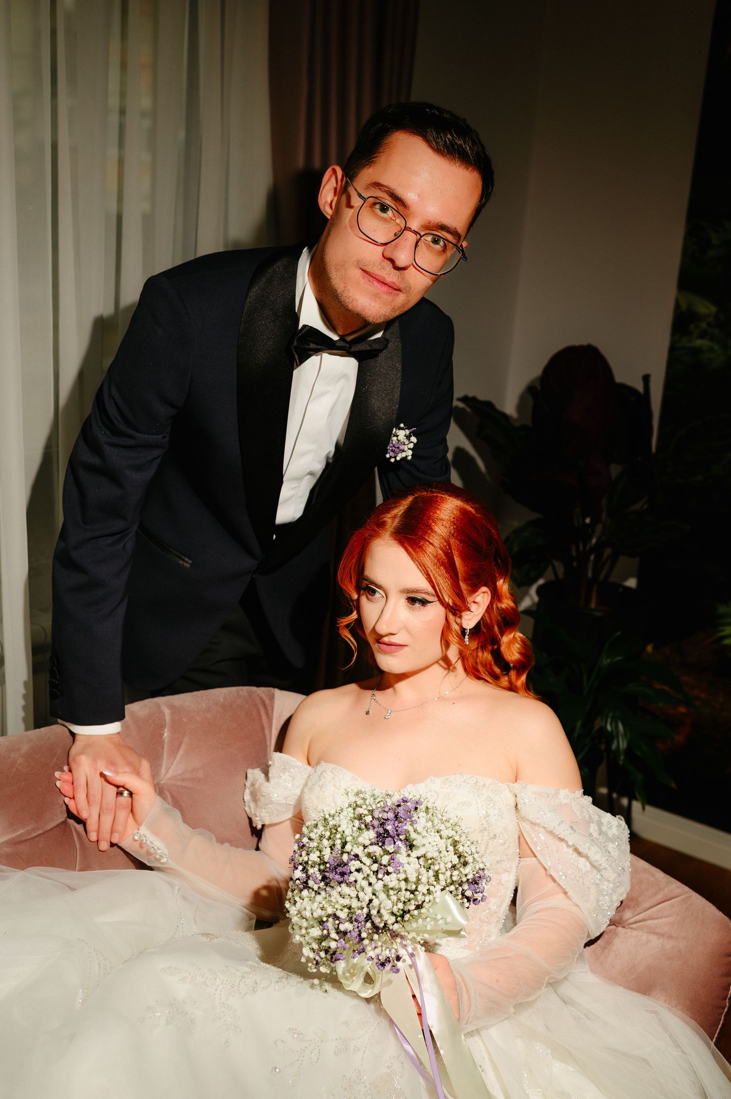 Elegant bride with red hair holding floral bouquet sits as groom in black tuxedo stands behind her.