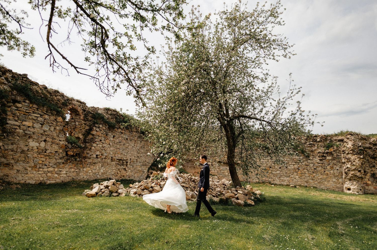 Bride and groom walking on green grass near rustic stone wall with blooming tree at outdoor wedding venue.