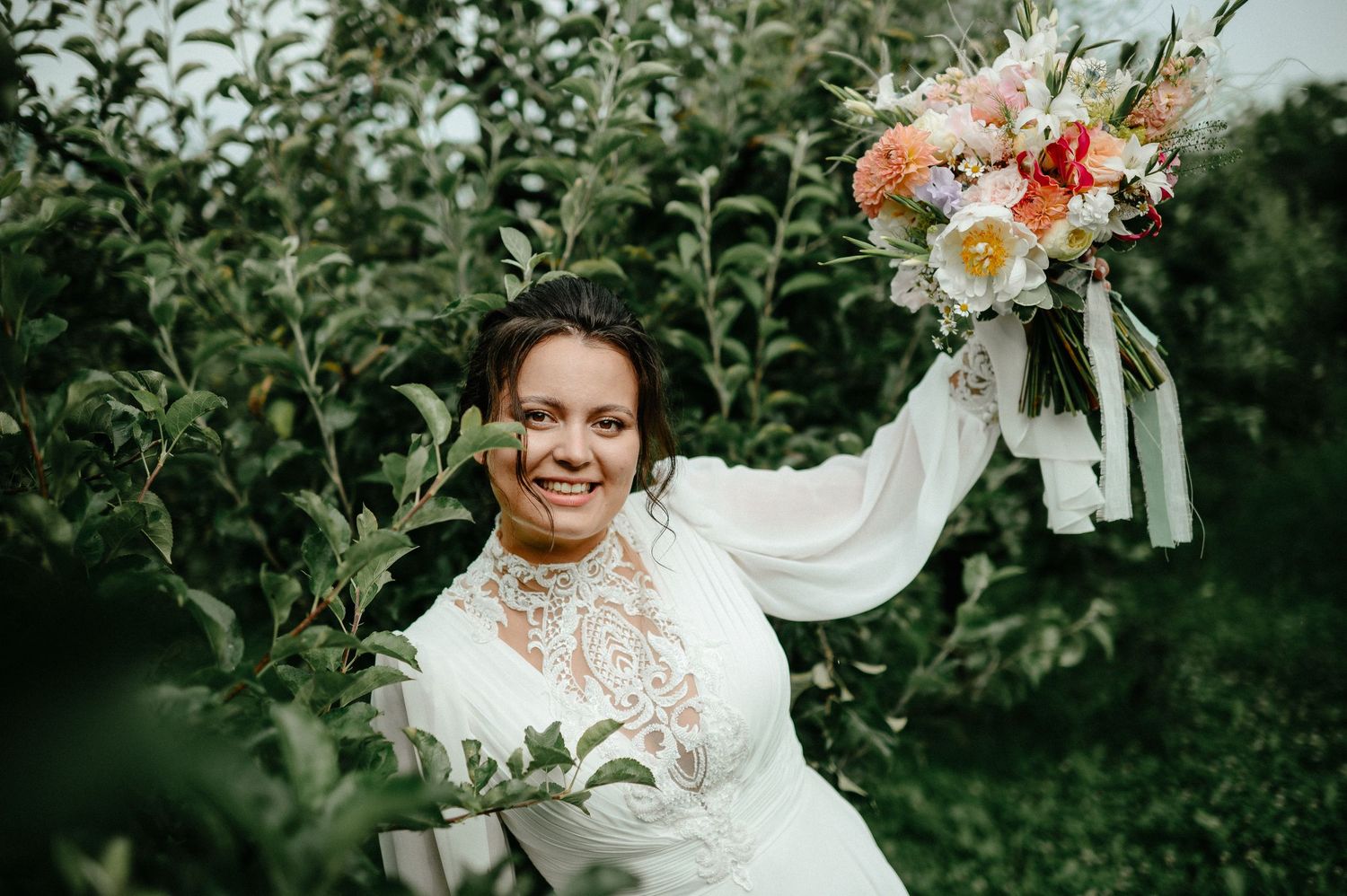 Bride in lace wedding gown holding colorful bouquet aloft in garden orchard.