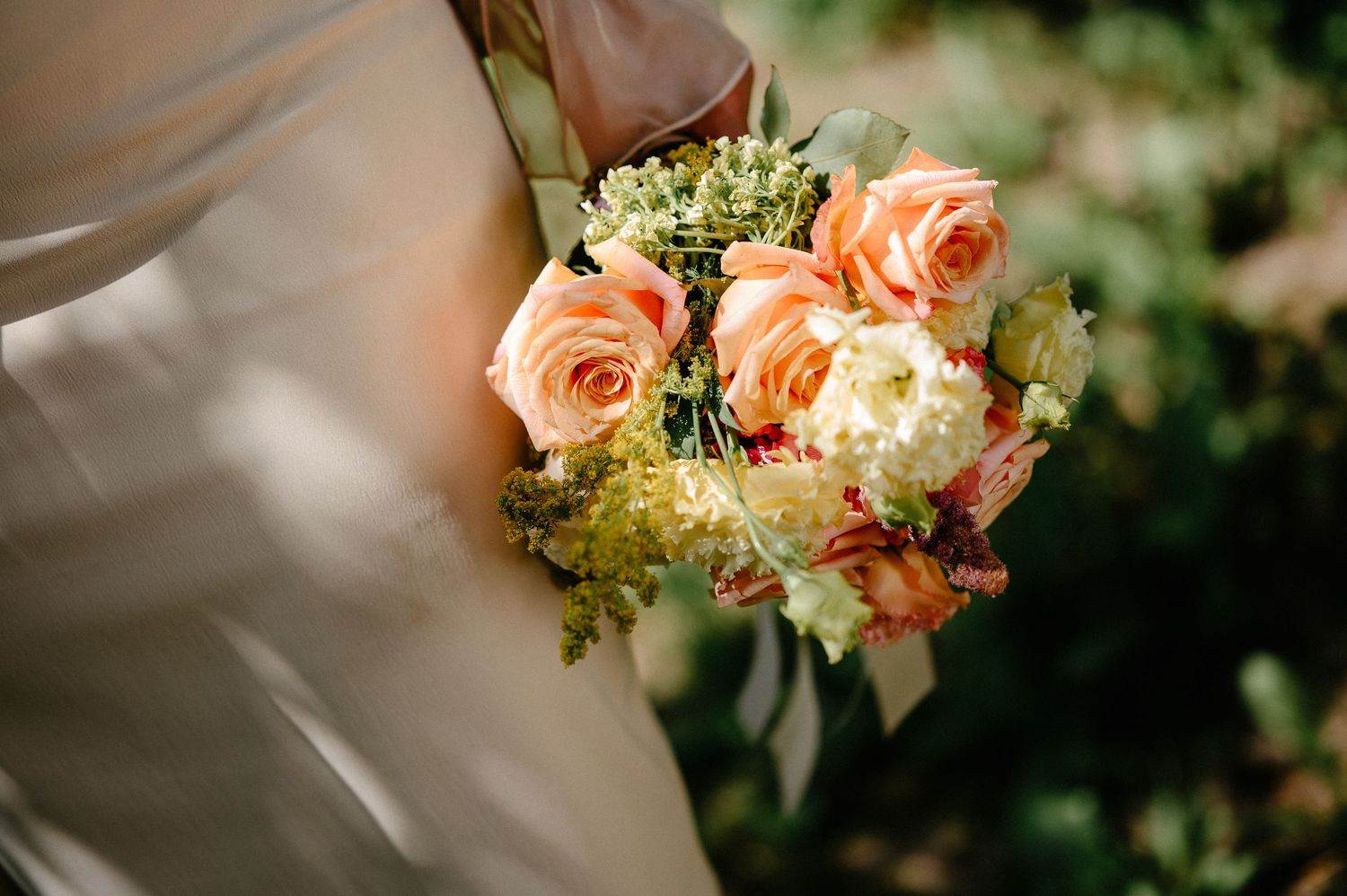 Bride holding a beautiful peach rose and white hydrangea wedding bouquet with greenery outdoors.