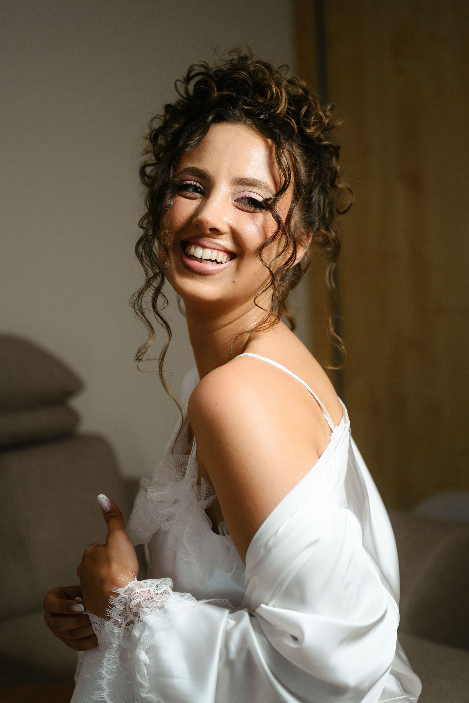 Smiling bride in white satin robe with elegant updo hairstyle and dramatic eye makeup, posing joyfully indoors.