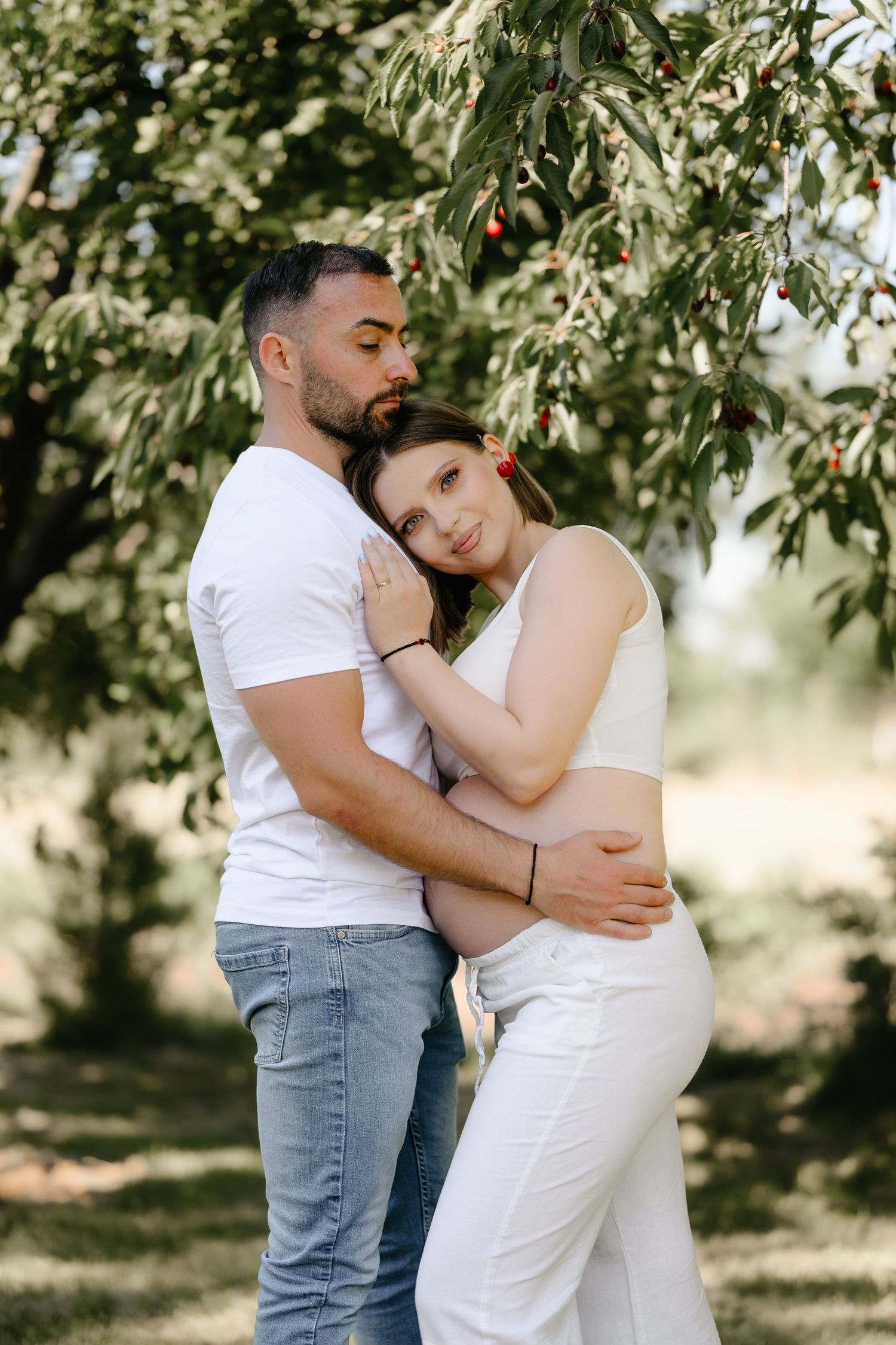 Couple embracing tenderly under cherry tree, pregnant woman in white outfit leaning on partner.