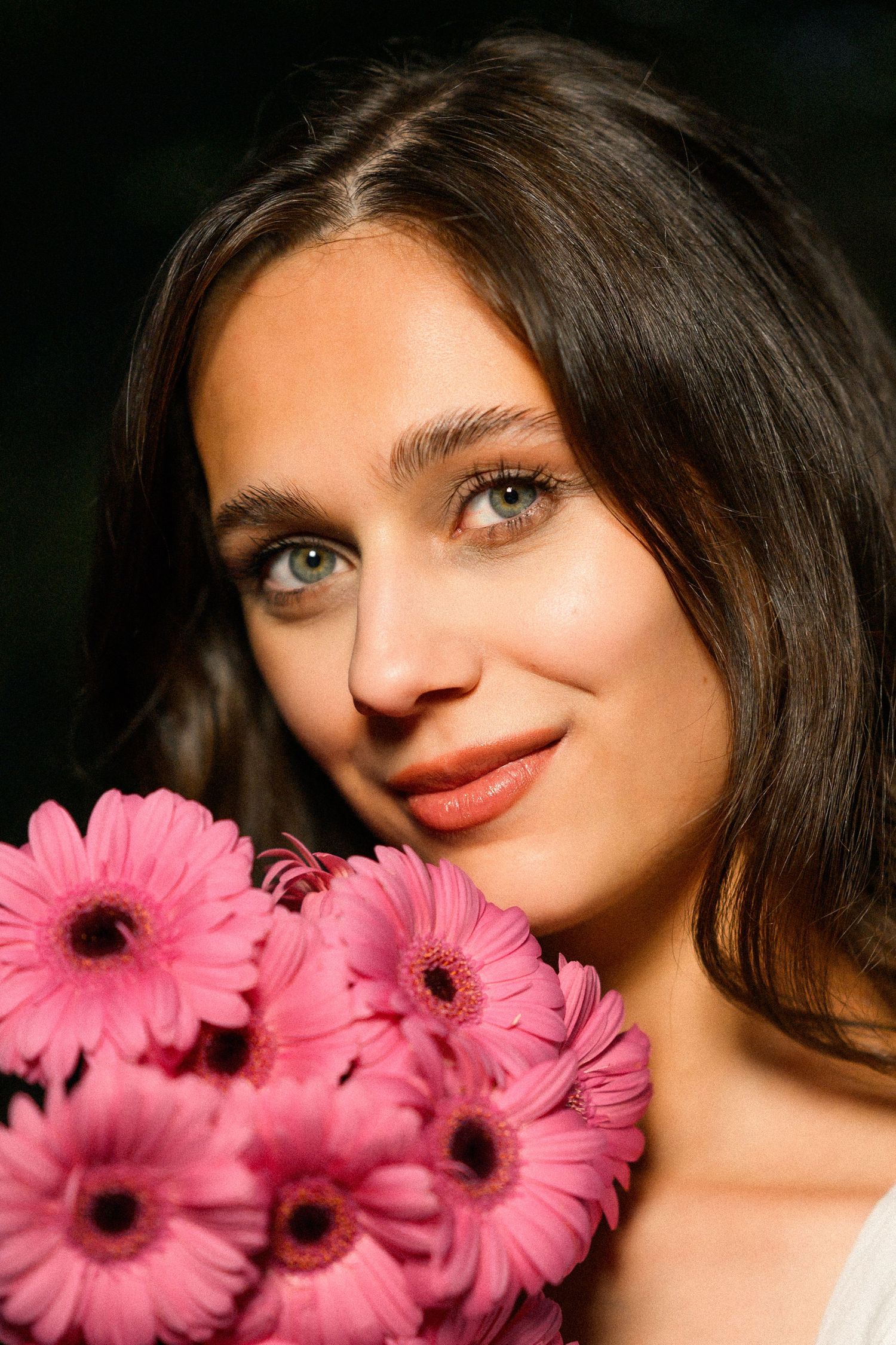 Young woman with green eyes holding a bouquet of pink gerbera daisies, smiling softly against a dark background.