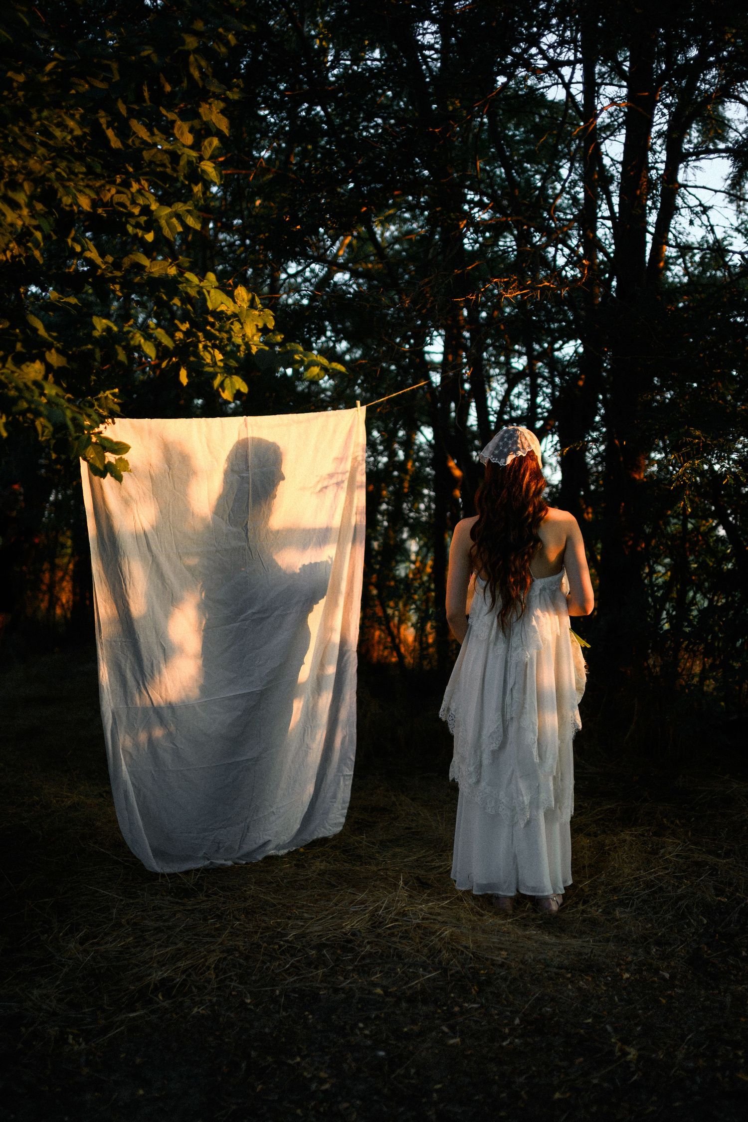 Woman in white dress holding a sheer fabric sheet with a shadowy figure silhouette in a dark forest at dusk.