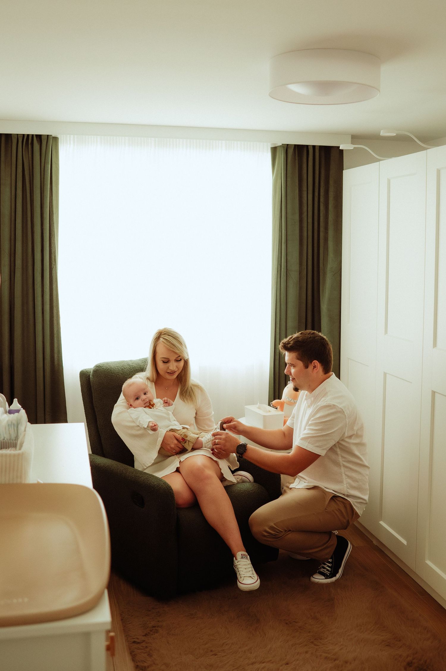 Young family with newborn in green nursery armchair, father kneeling beside mother and baby.
