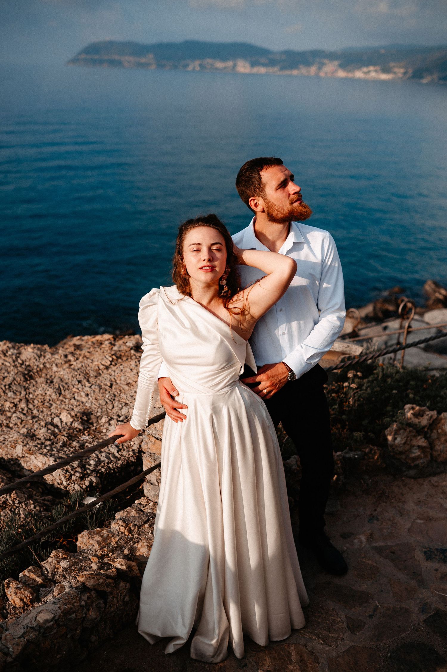 Couple in wedding attire embrace on stone terrace overlooking stunning blue Mediterranean sea at sunset.