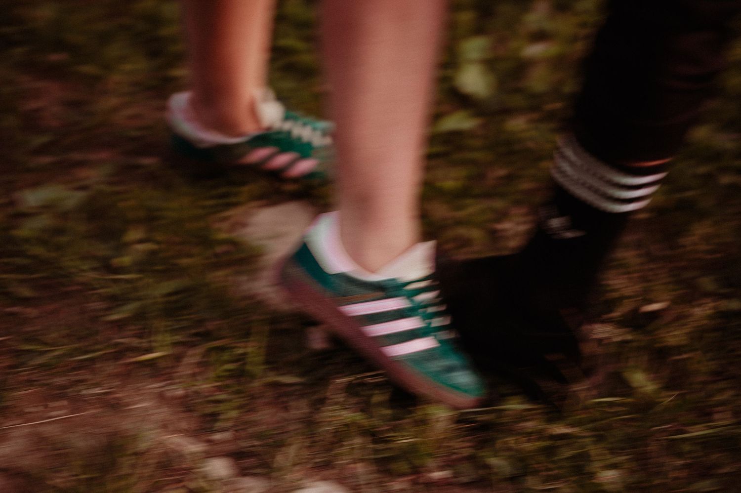 Blurred close-up of a woman wearing green and white Adidas sneakers standing outdoors on dark ground.