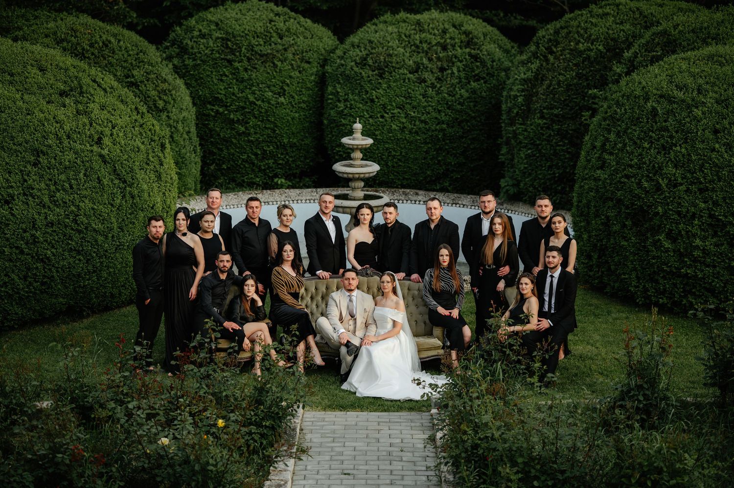 Wedding party group portrait in front of a stone fountain surrounded by sculpted green hedges.