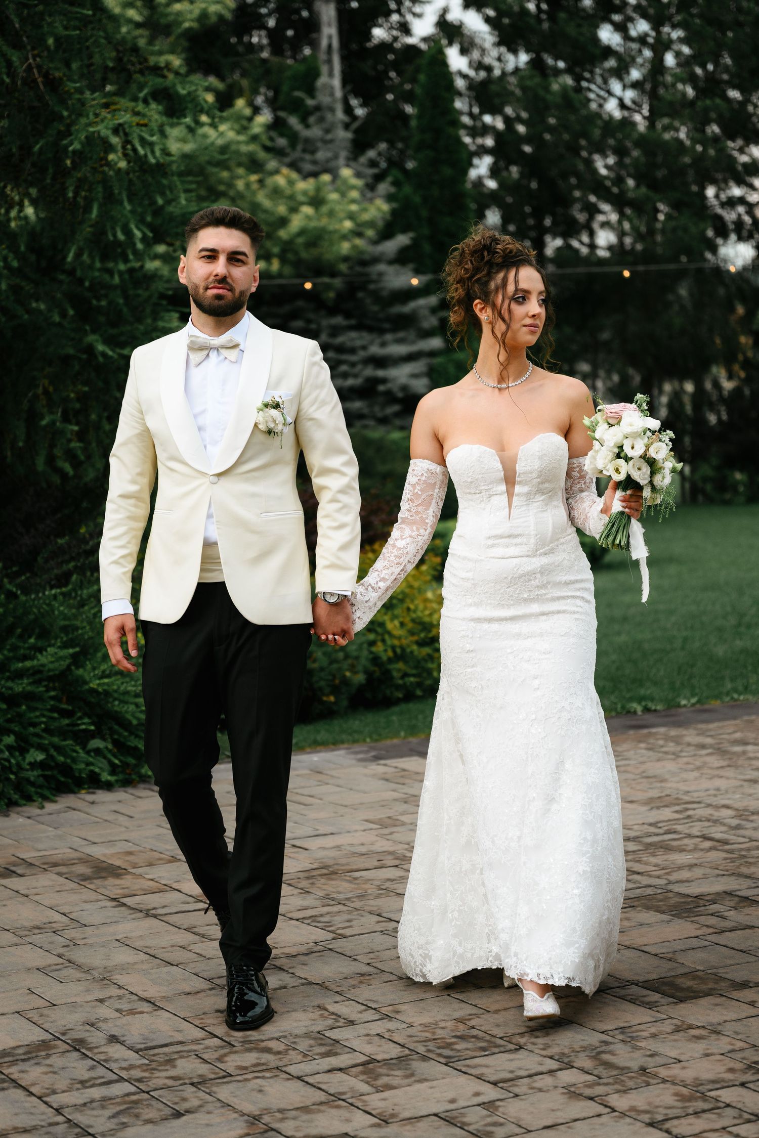 Bride and groom walking hand in hand outdoors, she in white lace gown with bouquet, he in ivory tuxedo jacket.