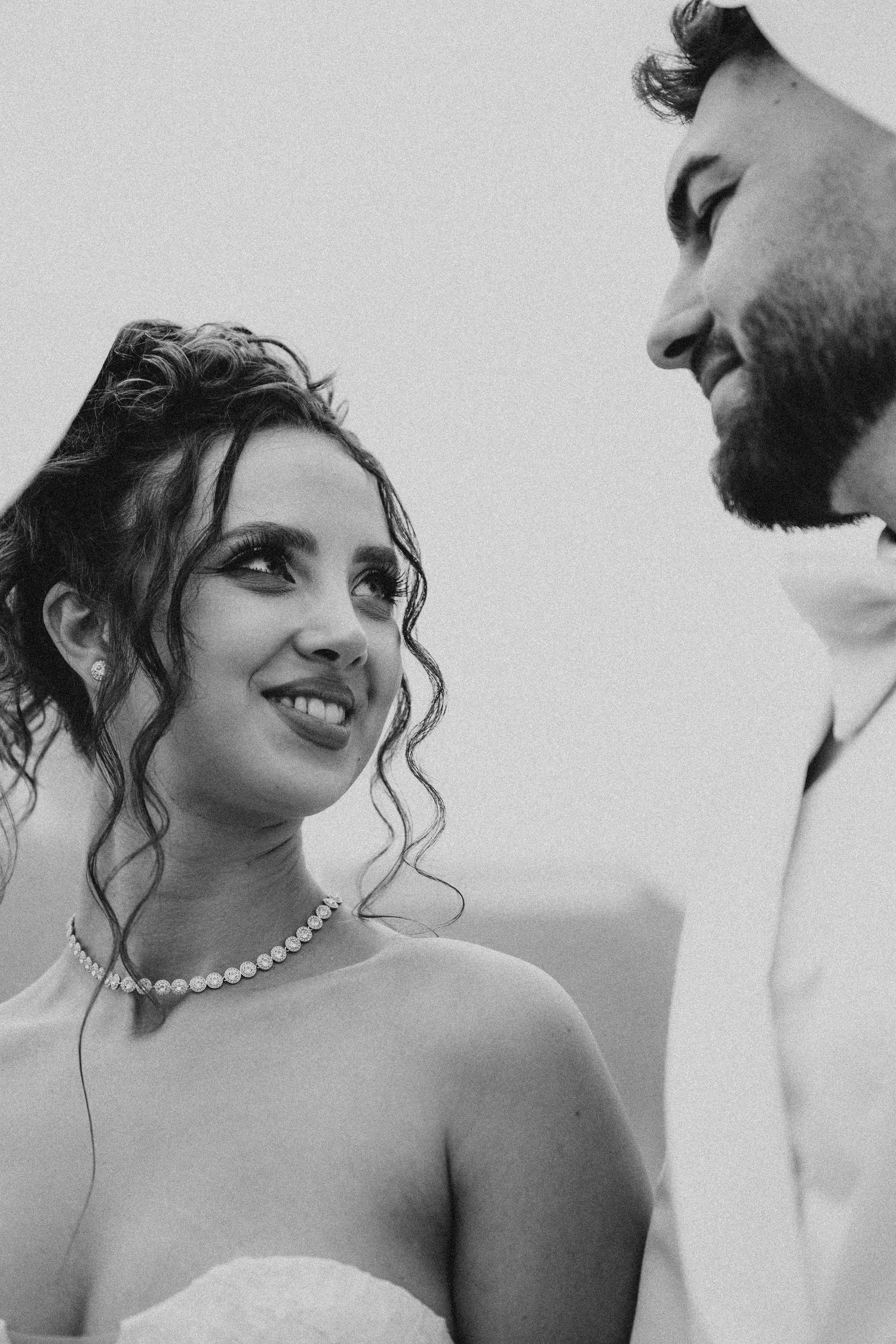 Black and white wedding photo of smiling bride wearing pearl necklace gazing at groom in white suit.
