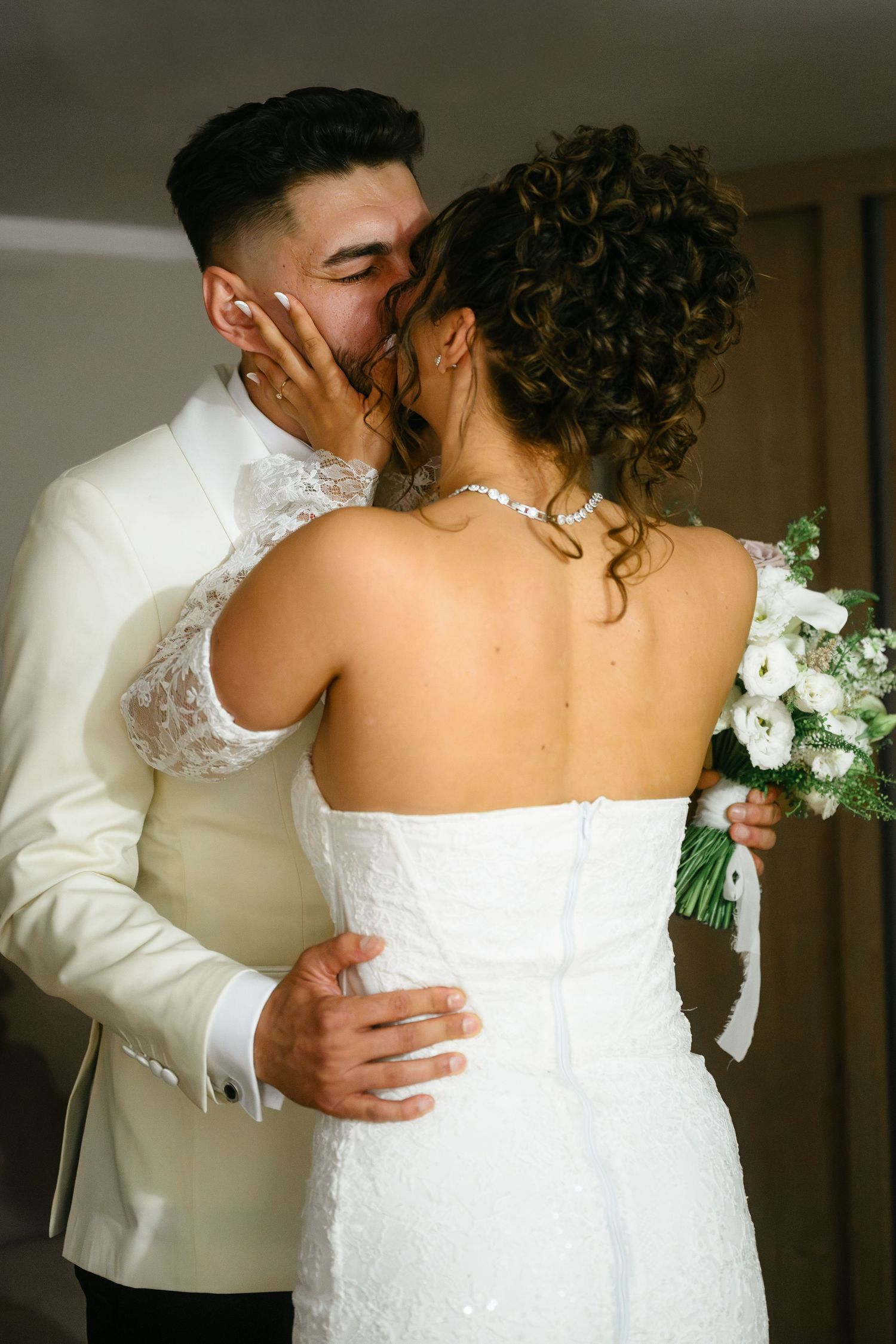 A bride and groom share a romantic kiss, she holds white flowers, wearing strapless gown as he embraces her in white suit.