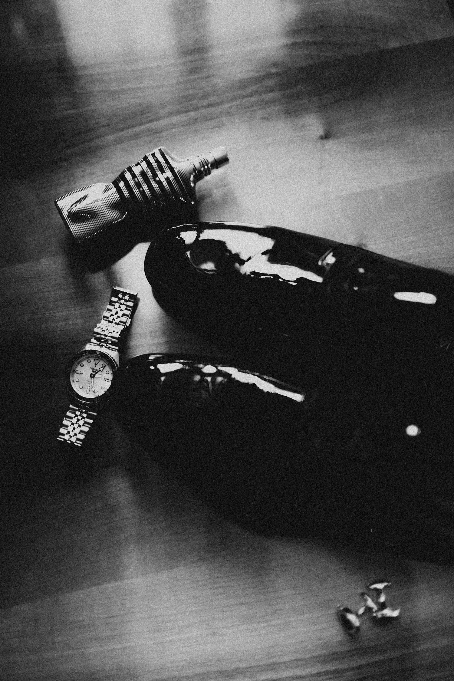 Black and white photo of mens dress shoes, watch, cufflinks, and cologne bottle on wooden floor.