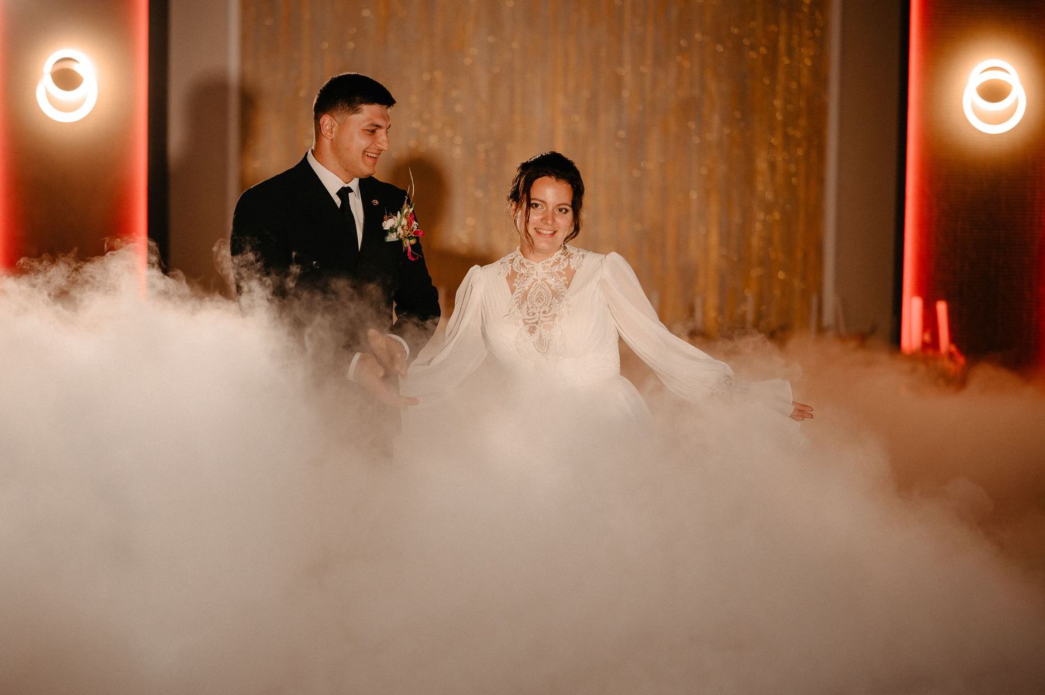 Bride and groom smile joyfully surrounded by thick fog machine clouds on elegant wedding reception stage.