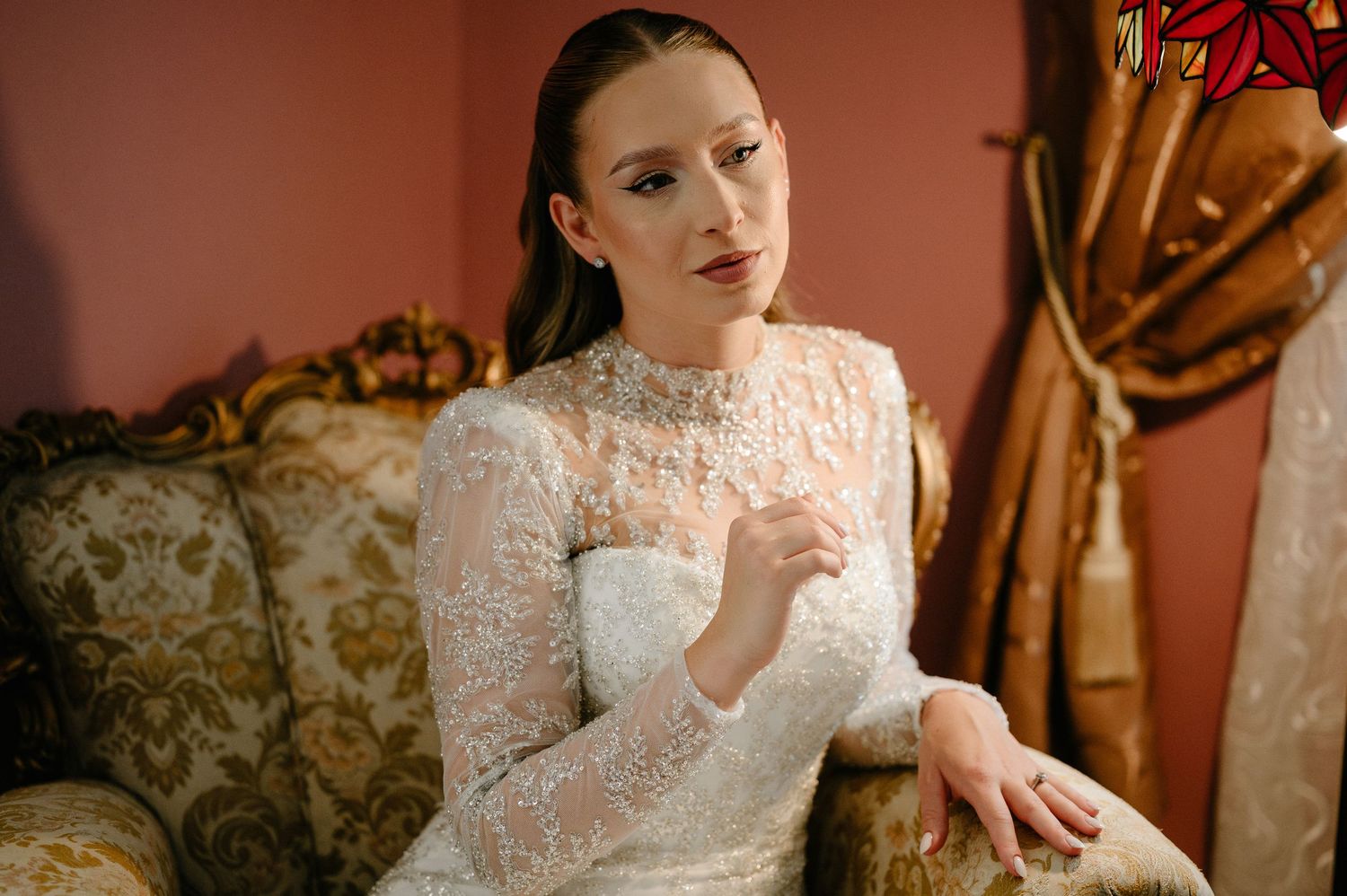 Graceful bride in vintage white lace high-neck gown seated on ornate gold sofa in pink room.
