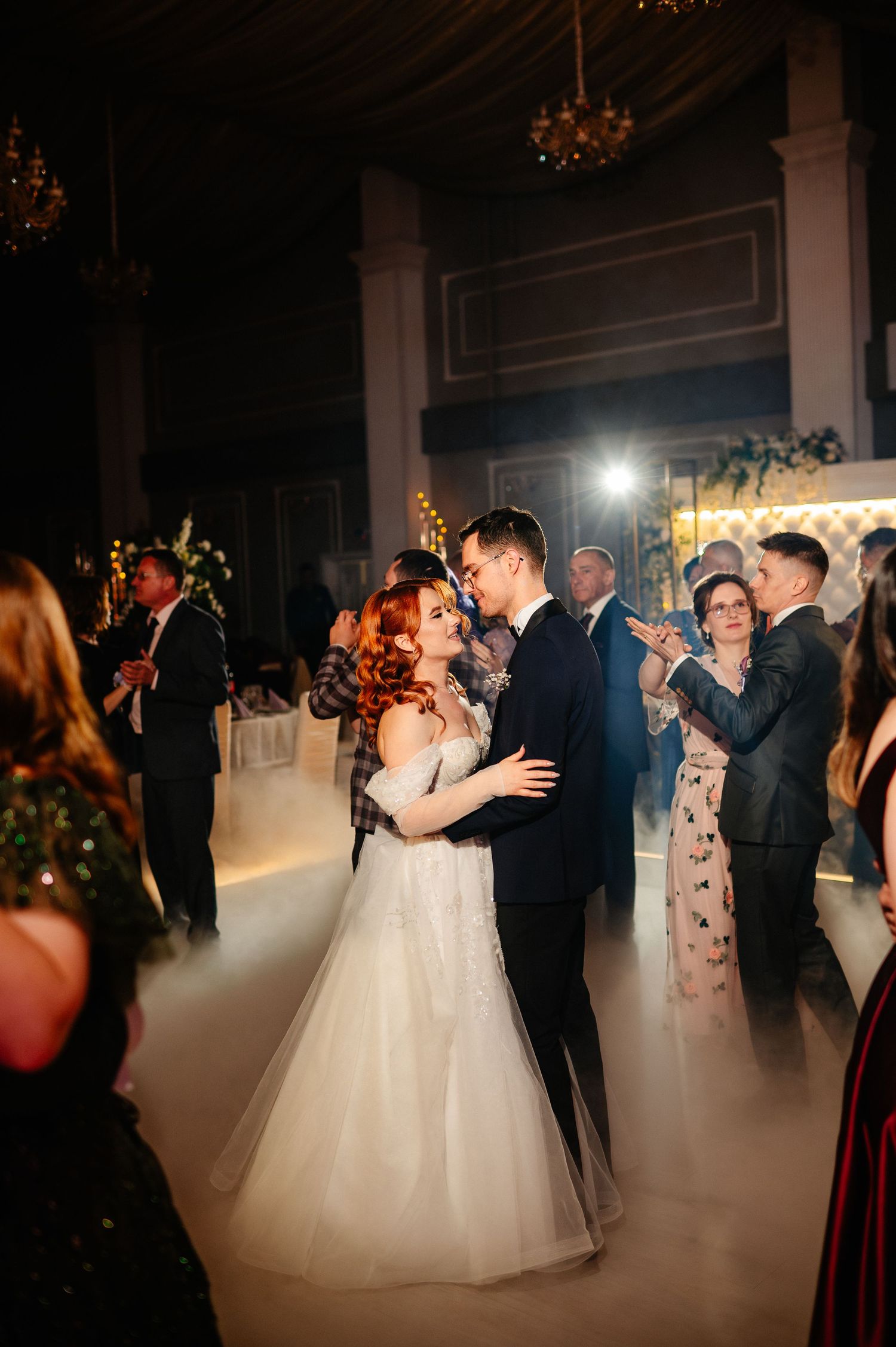Bride and groom share their first dance surrounded by guests in an elegant ballroom filled with soft fog lighting.