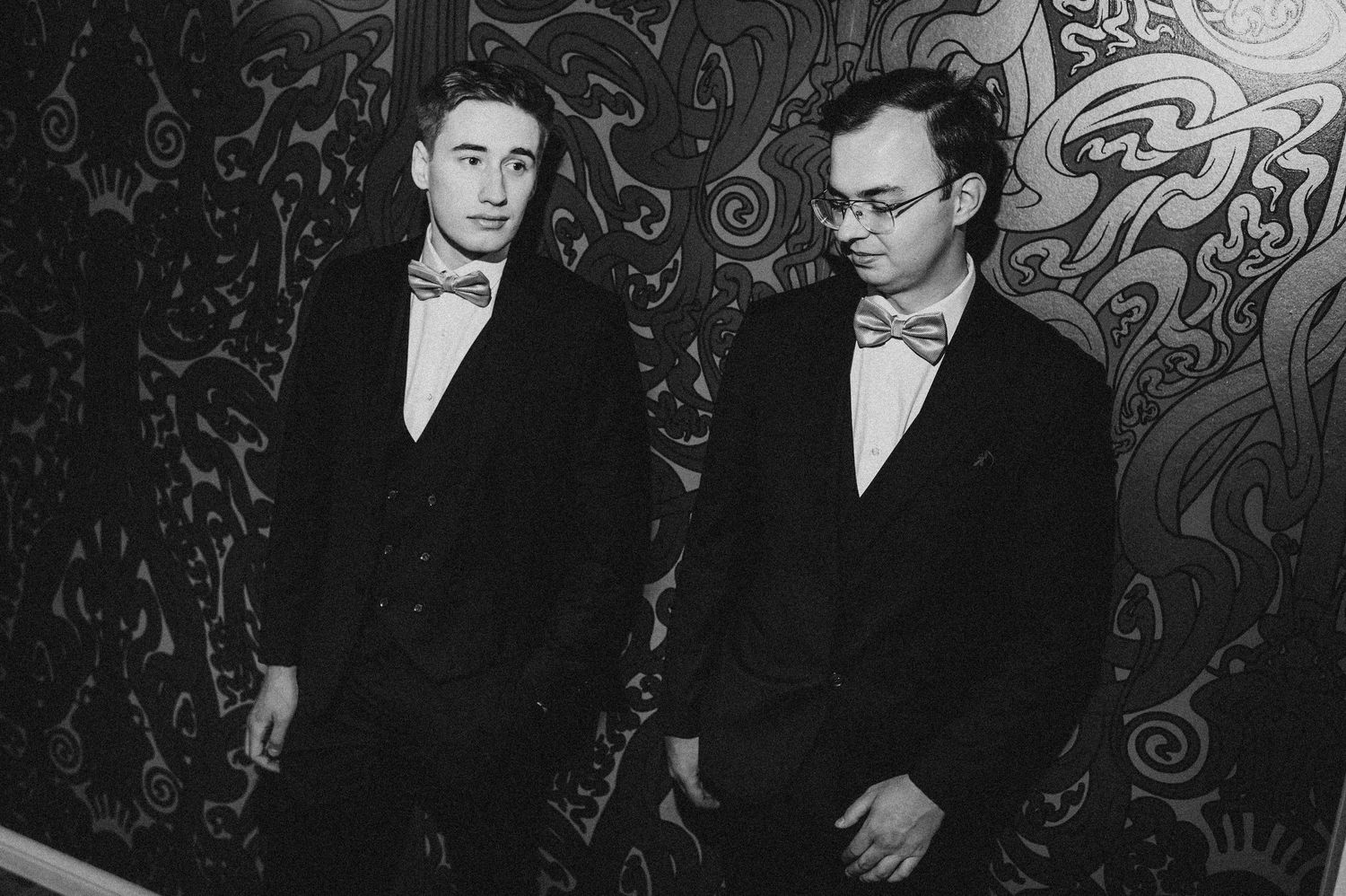 Two men in black tuxedos and bow ties pose formally in a black and white portrait photo.
