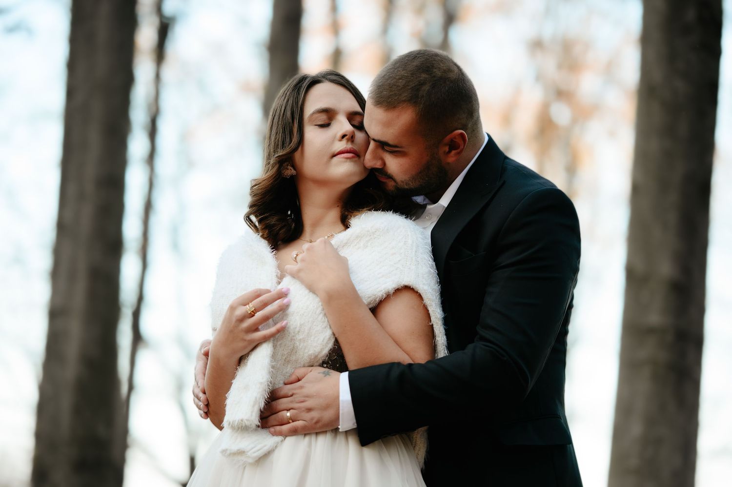 Romantic wedding couple embracing in a winter forest, bride in white fur wrap and groom in dark suit.