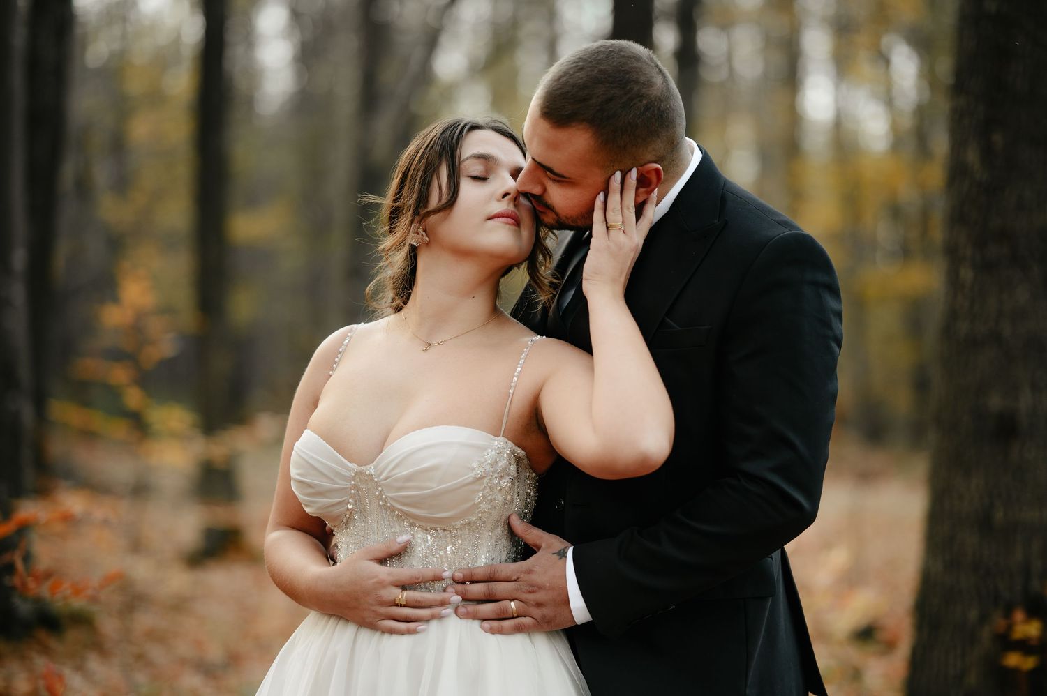 Bride and groom share a romantic moment in an autumn forest, with the groom in a black suit and bride in a white gown.
