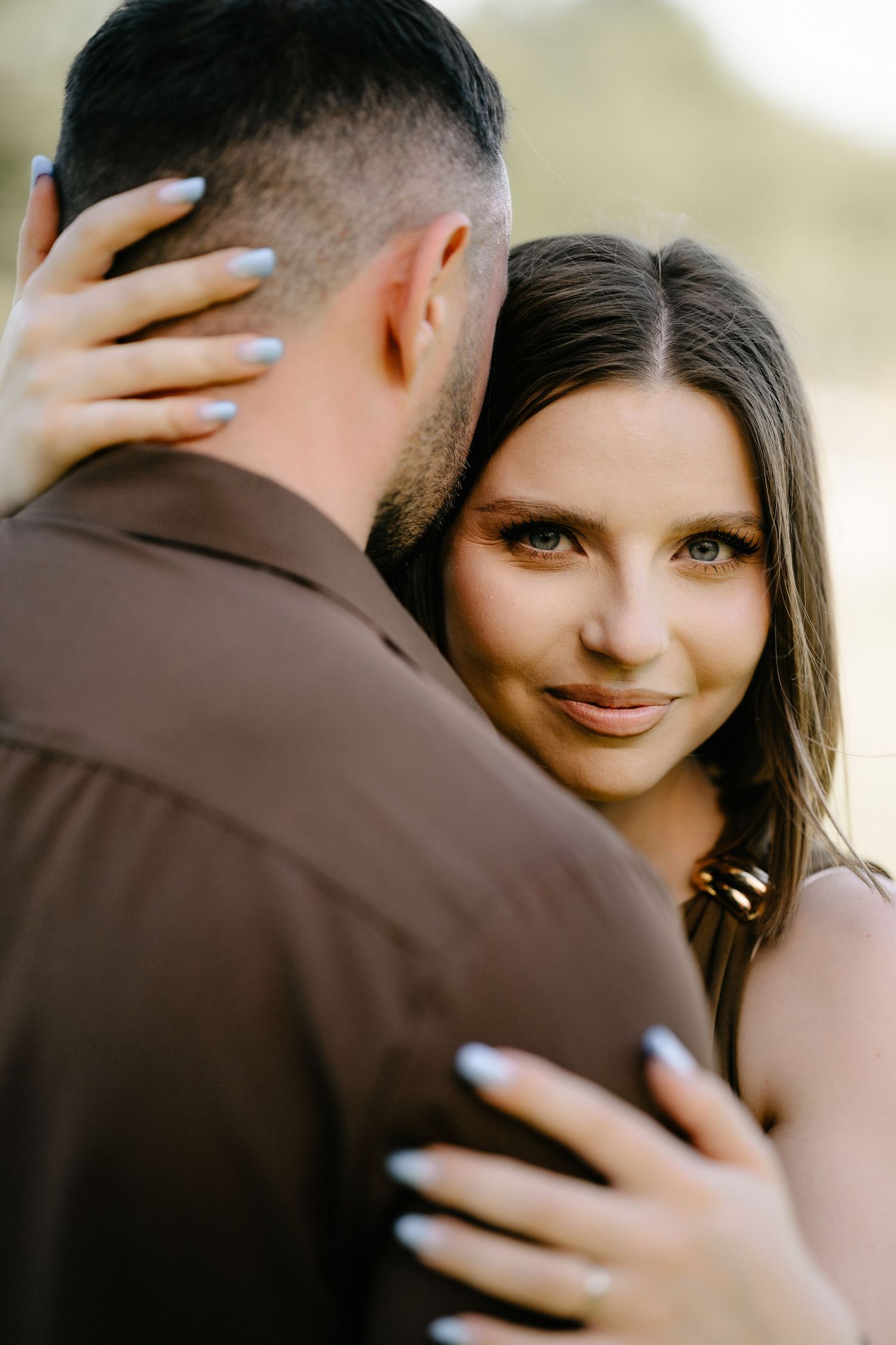 Woman with light nails embraces man in brown shirt, smiling warmly in soft outdoor light.