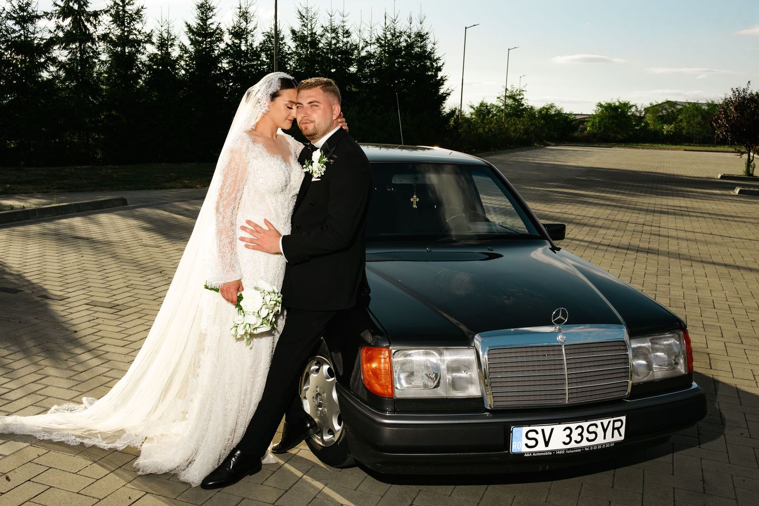 Bride and groom posing together beside a black Mercedes-Benz sedan on their wedding day outdoors.
