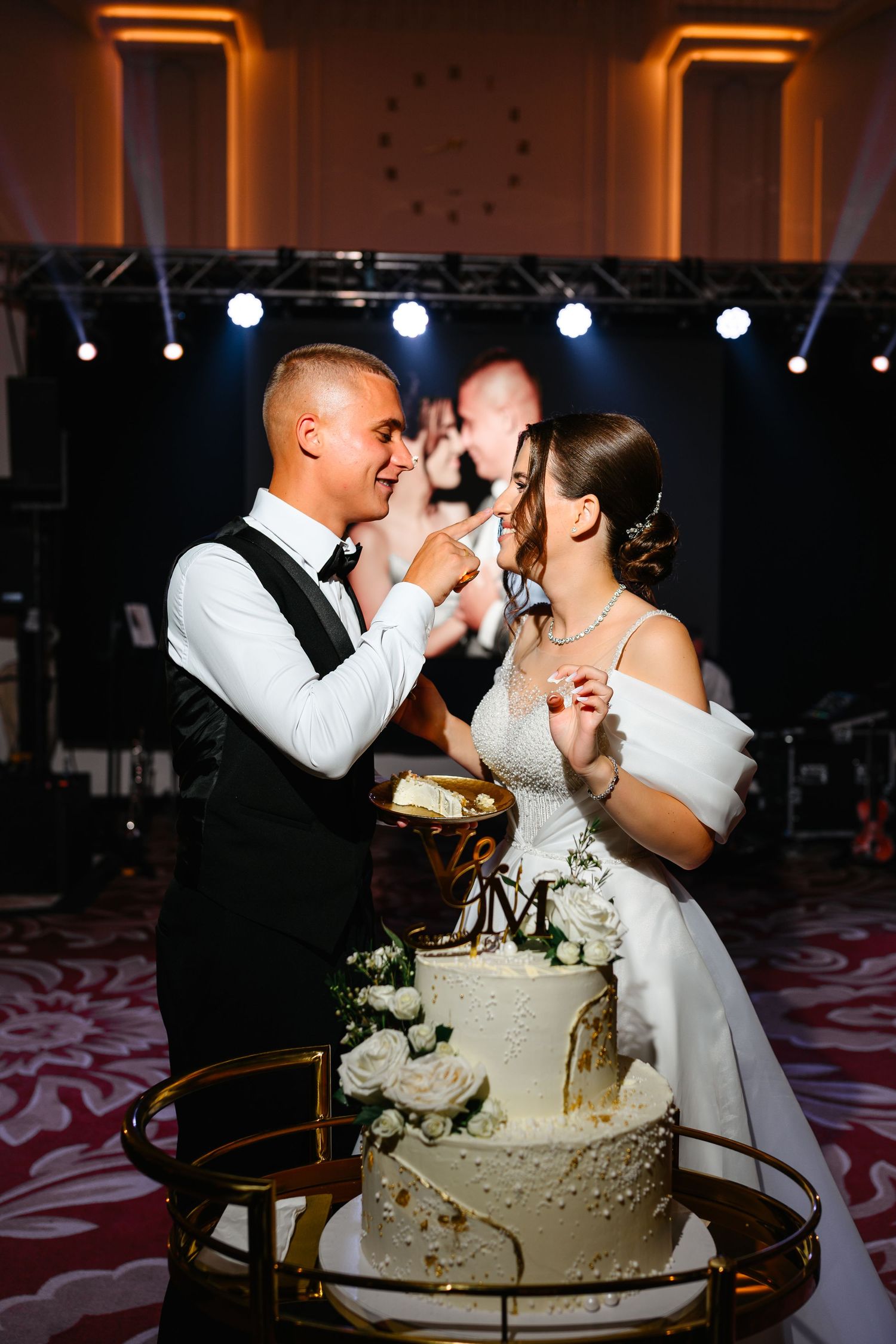 Bride and groom feeding each other wedding cake at elegant reception venue with stage lights.