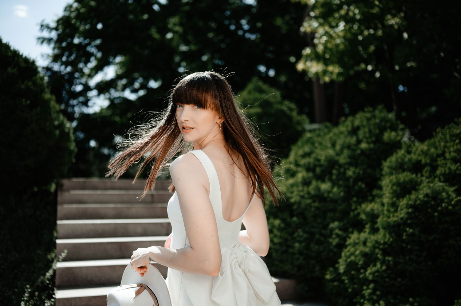Woman in white backless dress with flowing hair looking over shoulder on garden stairs.