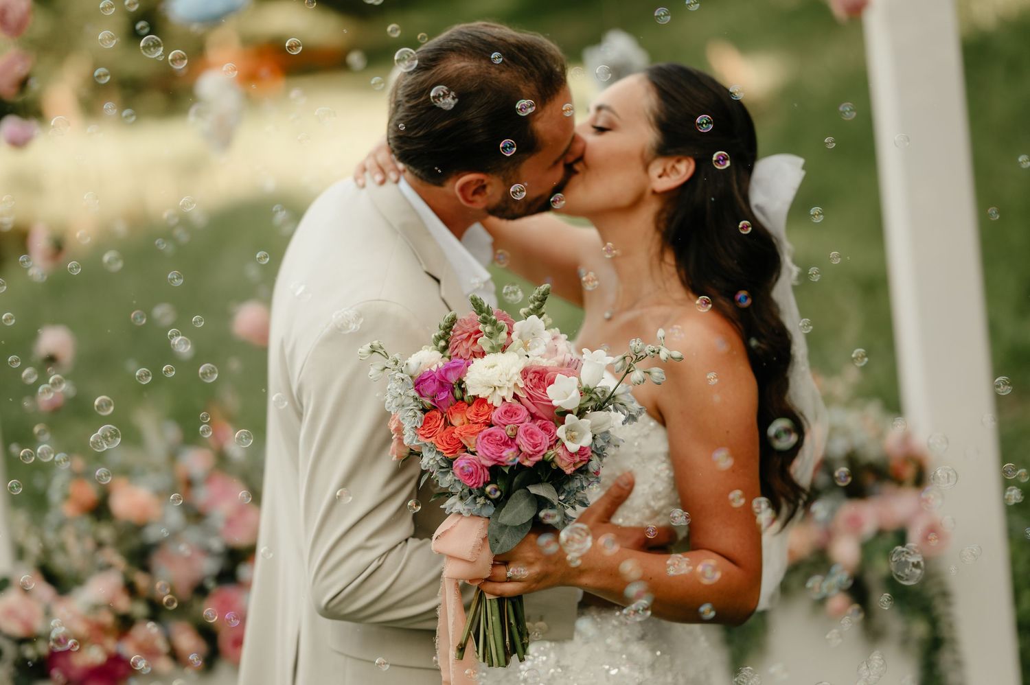 Newlyweds share a romantic kiss surrounded by floating bubbles at their outdoor wedding ceremony.