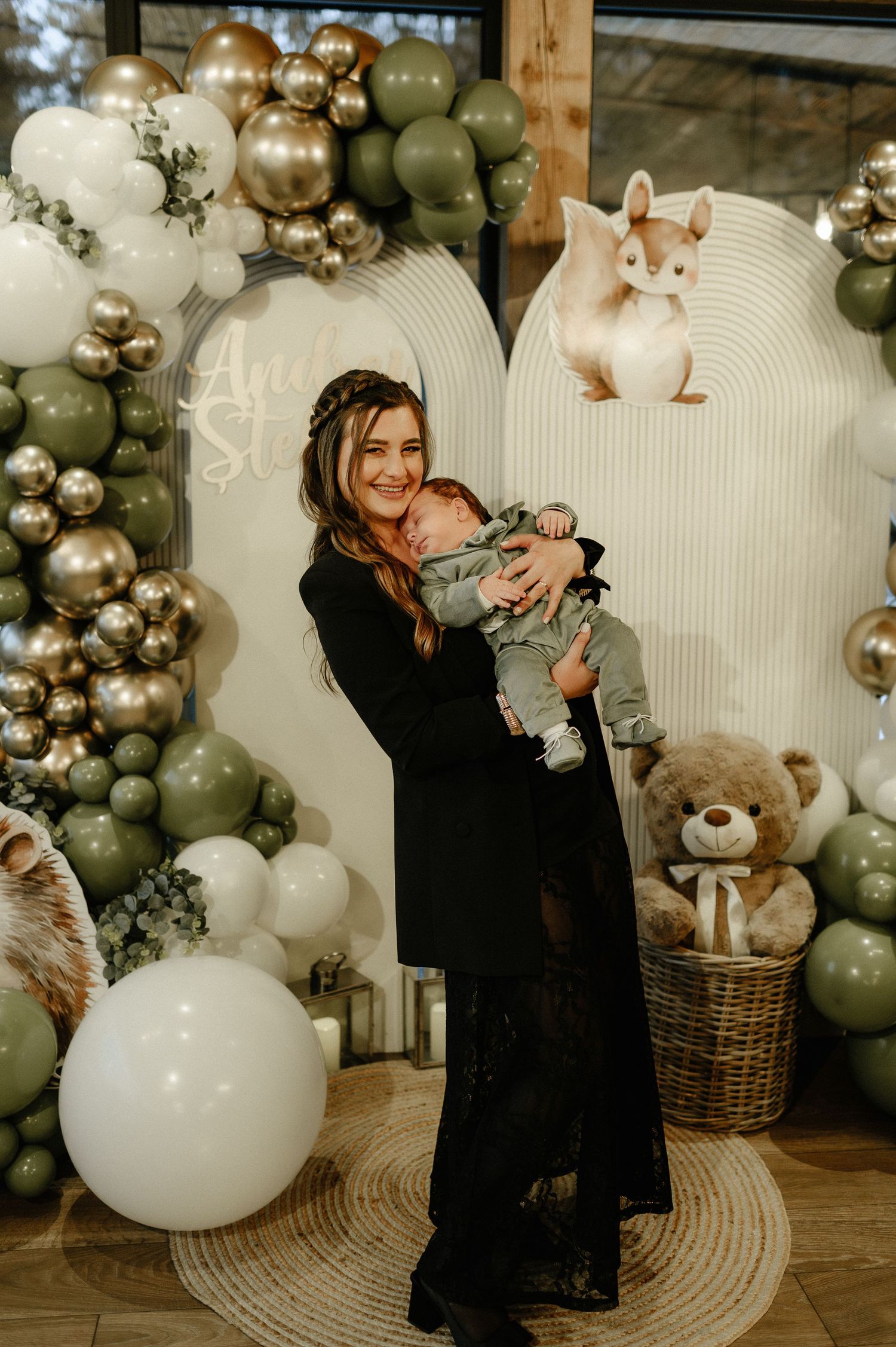 Woman in black dress holding newborn baby at elegant sage green and gold balloon decorated baby shower.
