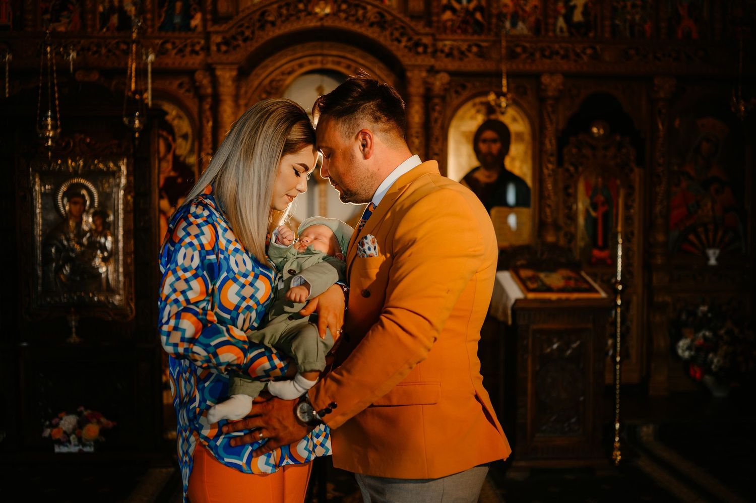Young couple gazing at each other while holding newborn in richly decorated Orthodox church.
