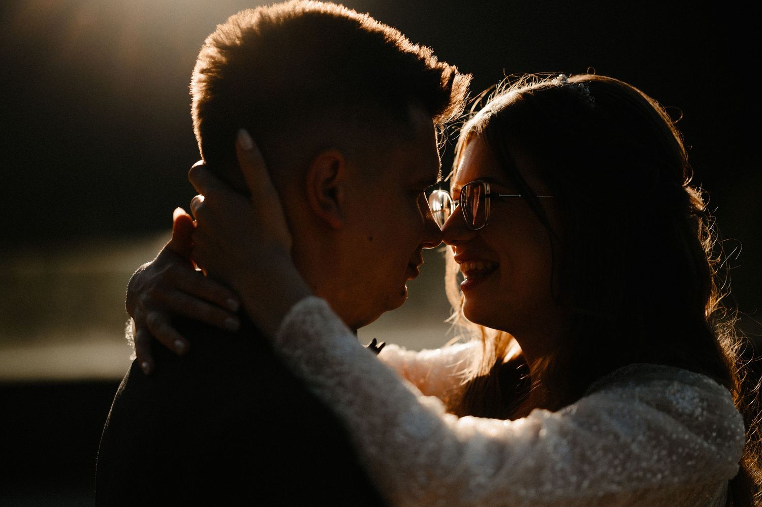 Bride and groom embracing closely in dramatic dark romantic lighting outdoors.