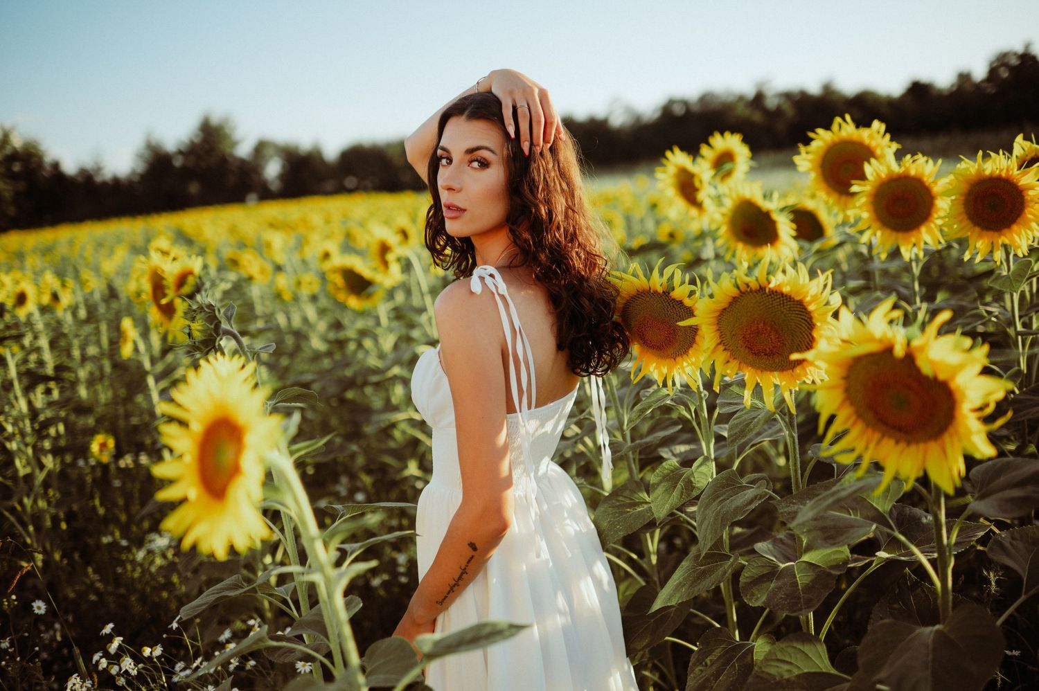 Brunette woman in white dress and straw hat gazing over shoulder in a sunflower field at sunset.