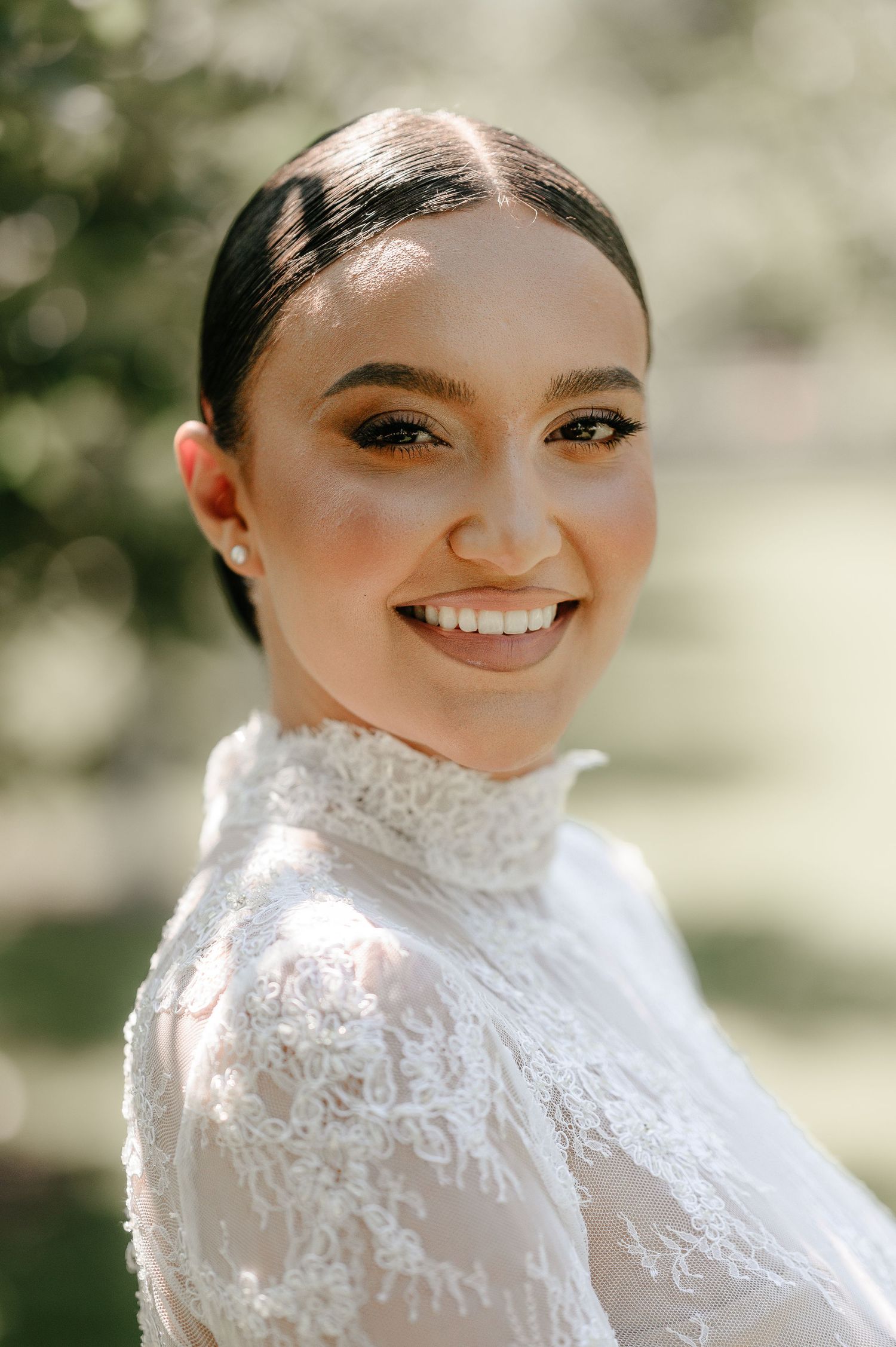 Close-up portrait of a smiling bride with sleek updo wearing a high-neck lace wedding dress outdoors.