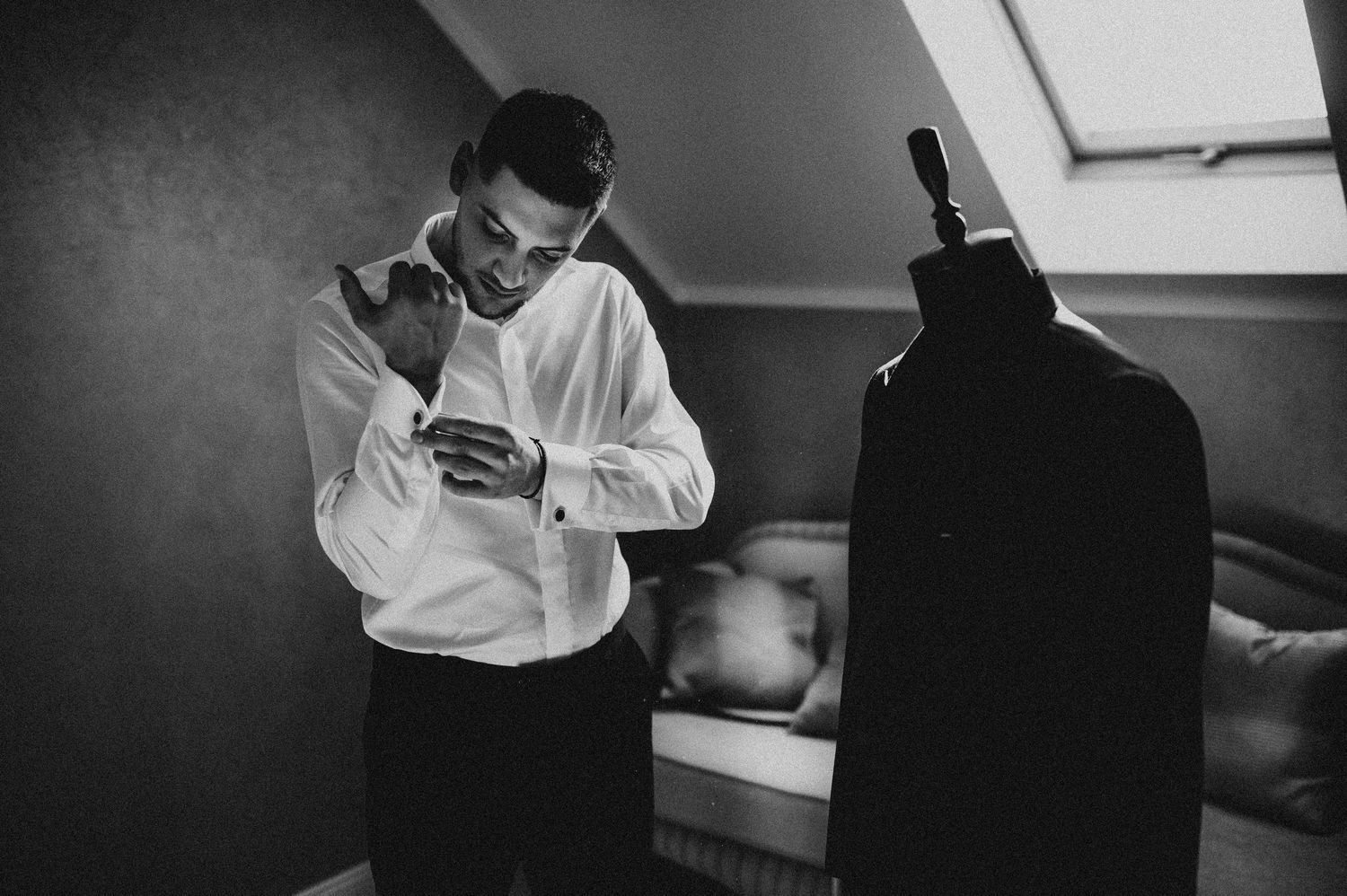Black and white photo of groom adjusting cufflinks near suit on mannequin.