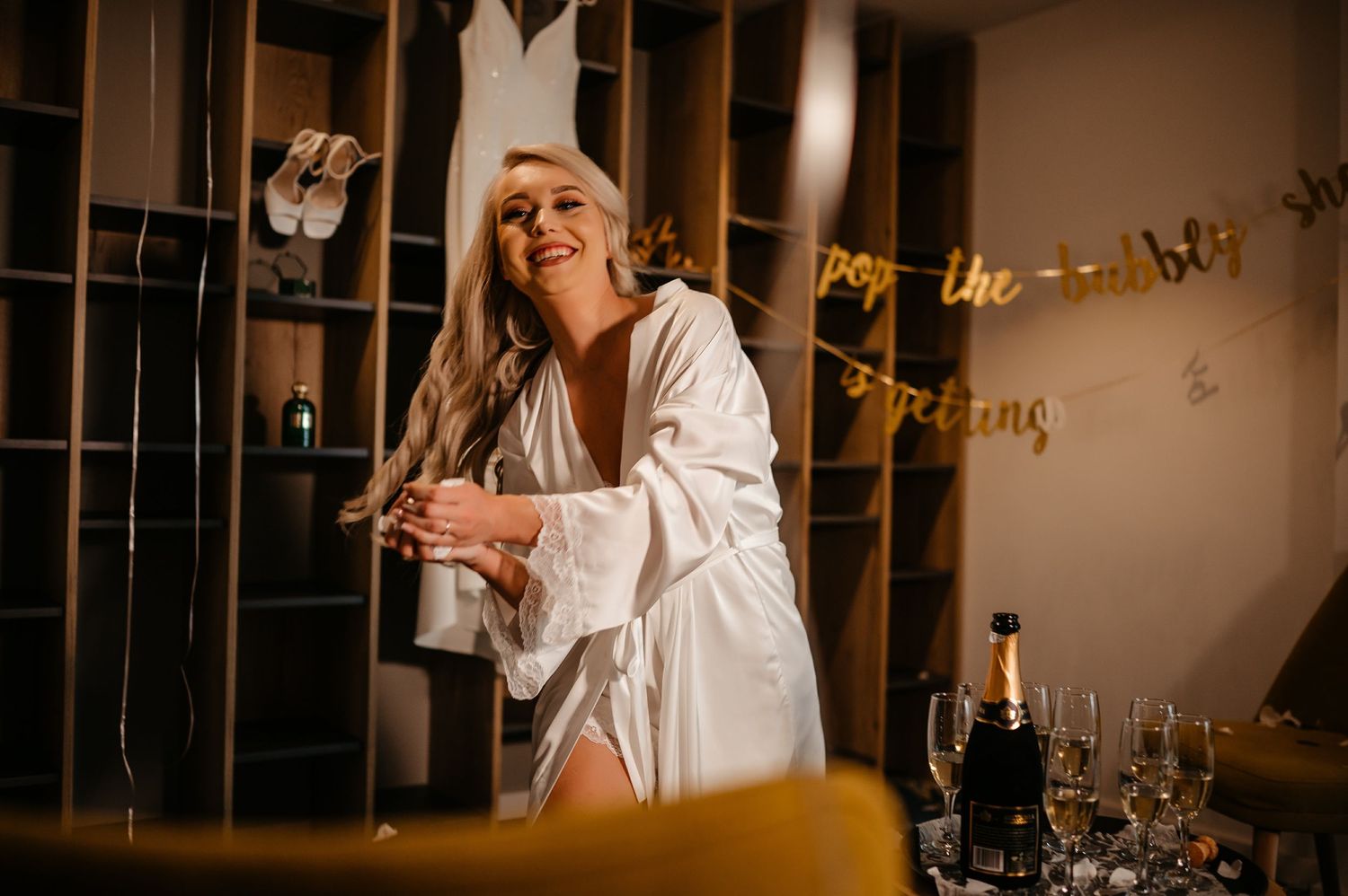 Smiling bride in white satin robe celebrating with champagne near gold banner decorations before wedding.