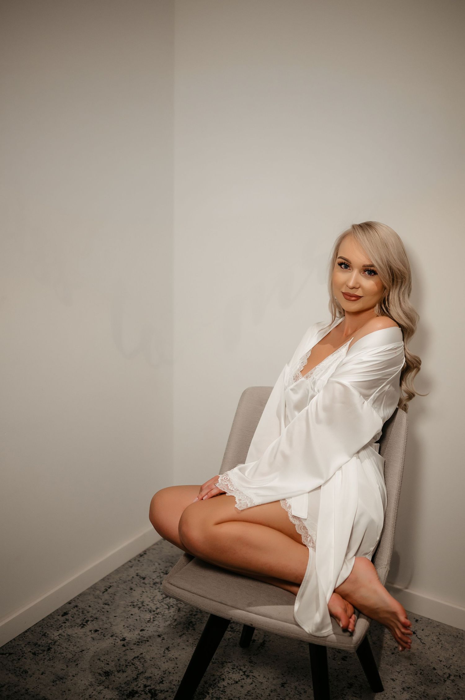 Blonde bride posing elegantly in a white satin robe seated on a chair during bridal preparations.