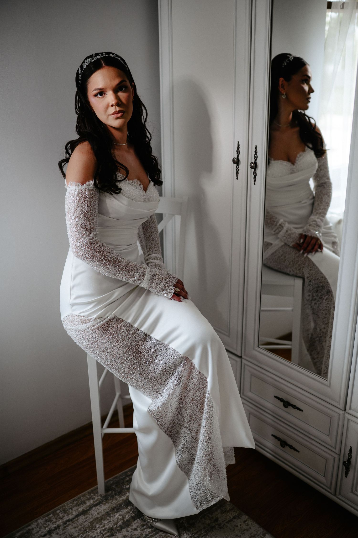 Elegant bride in off-shoulder lace wedding gown sitting beside a wardrobe mirror before wedding.