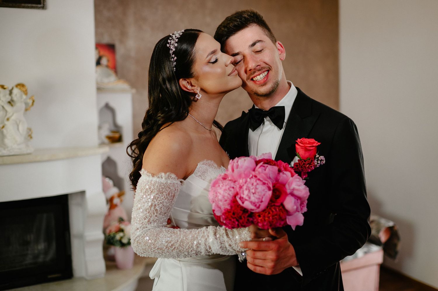 Bride kisses groom on cheek as he smiles broadly, both holding bright pink peony wedding bouquet.