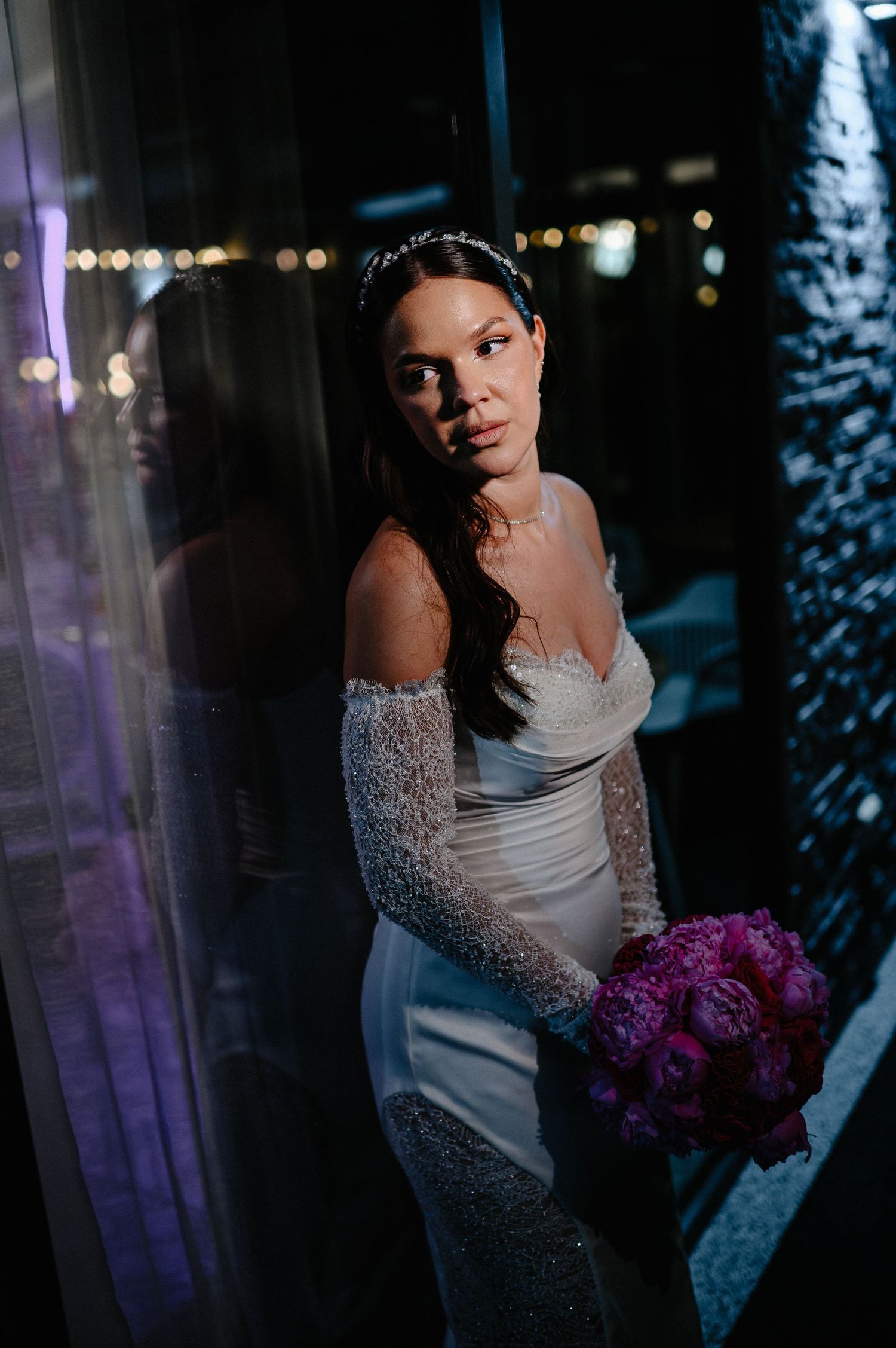 Elegant bride in off-shoulder lace wedding gown holding pink peony bouquet posing at night venue.