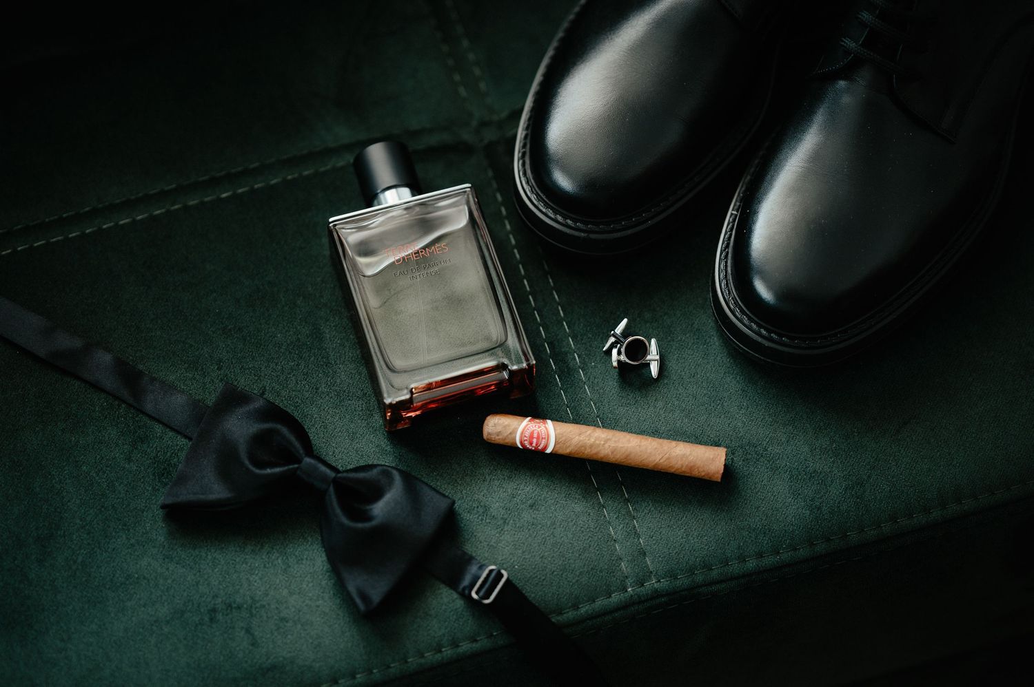 Wedding groom accessories flat lay with cologne, cigar, bow tie, cufflinks, and black dress shoes.