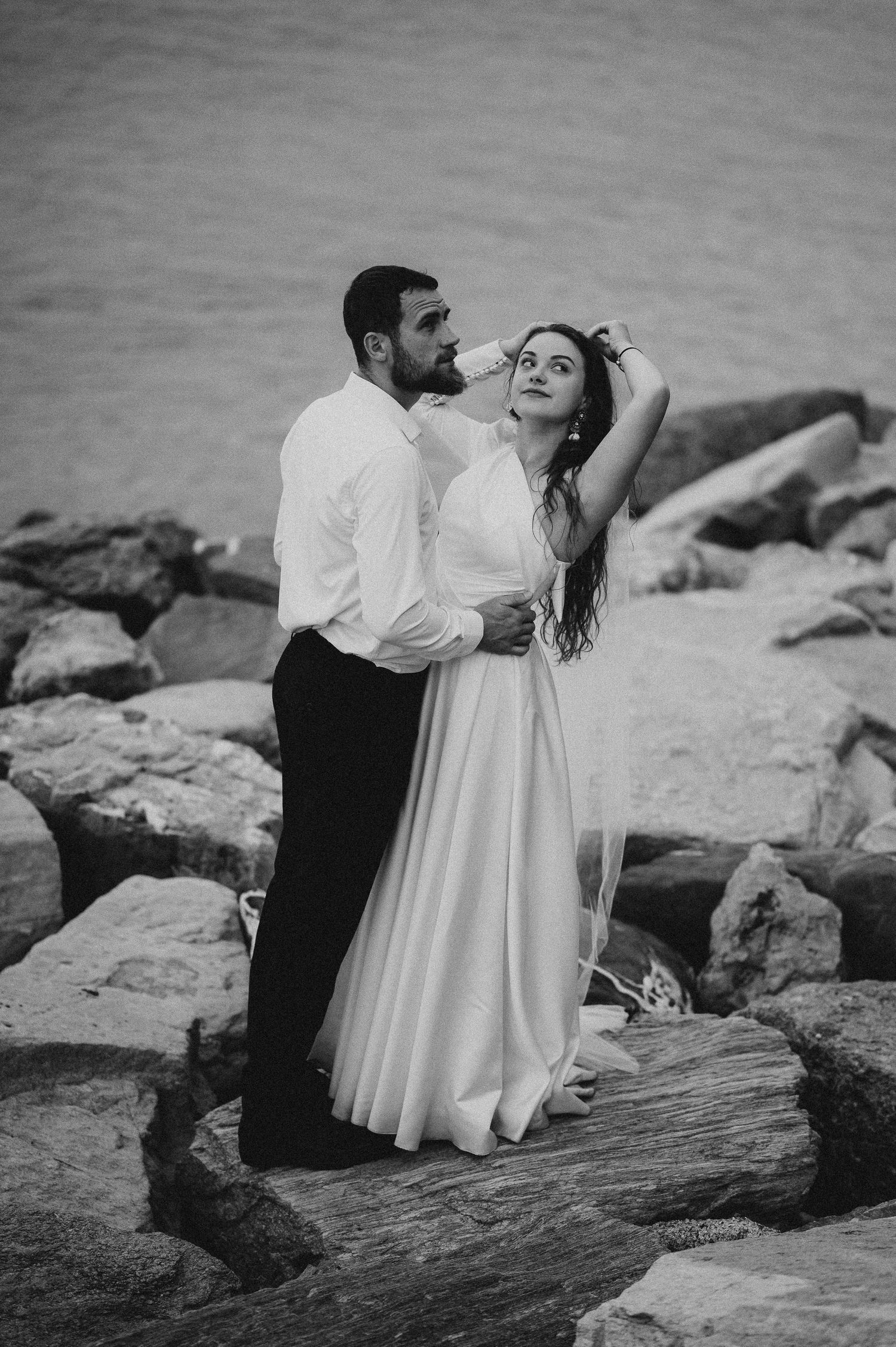 Black and white photo of couple embracing on rocky seaside in wedding attire.
