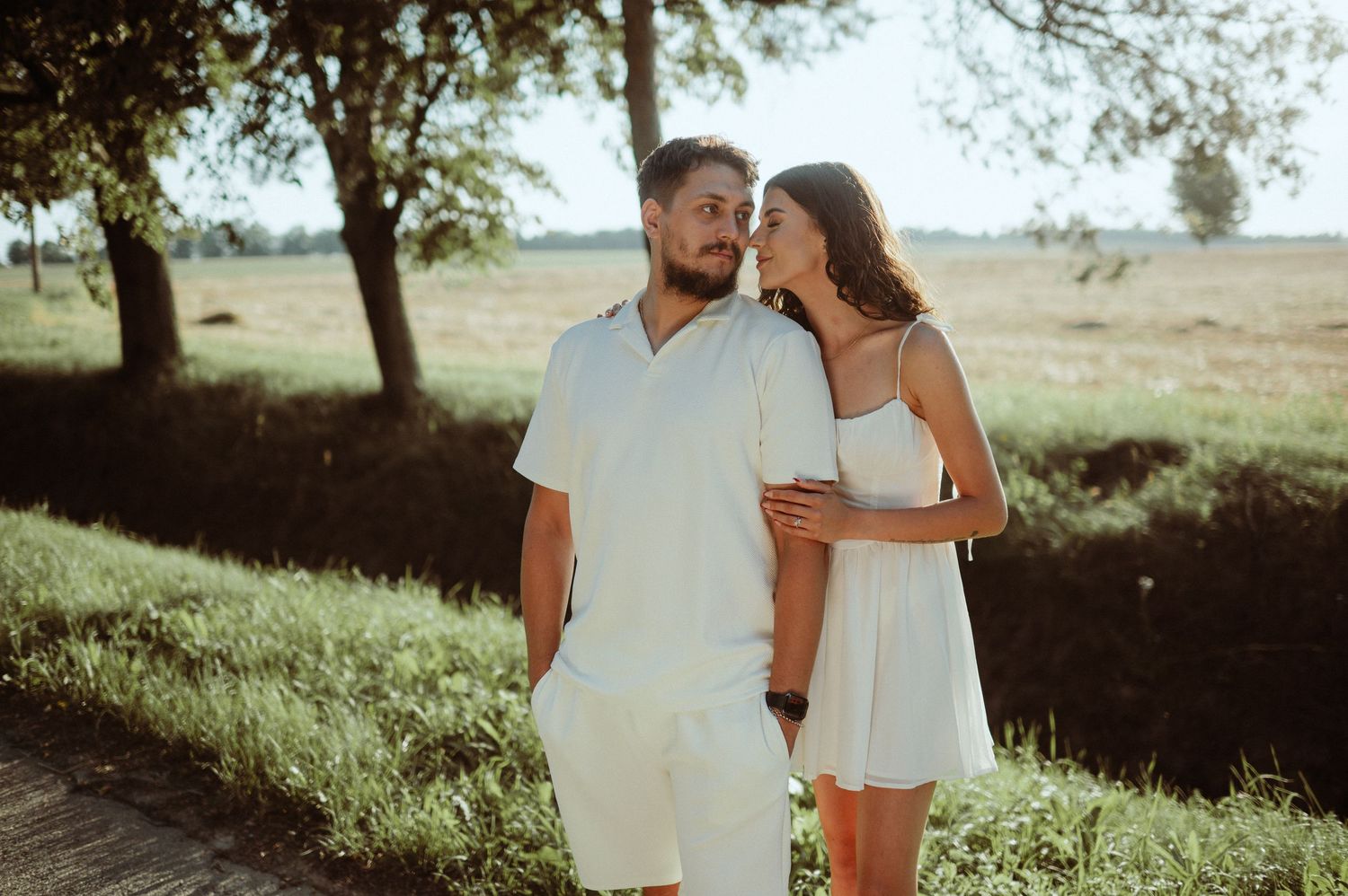 Couple in white outfits embracing near trees in golden sunlight.
