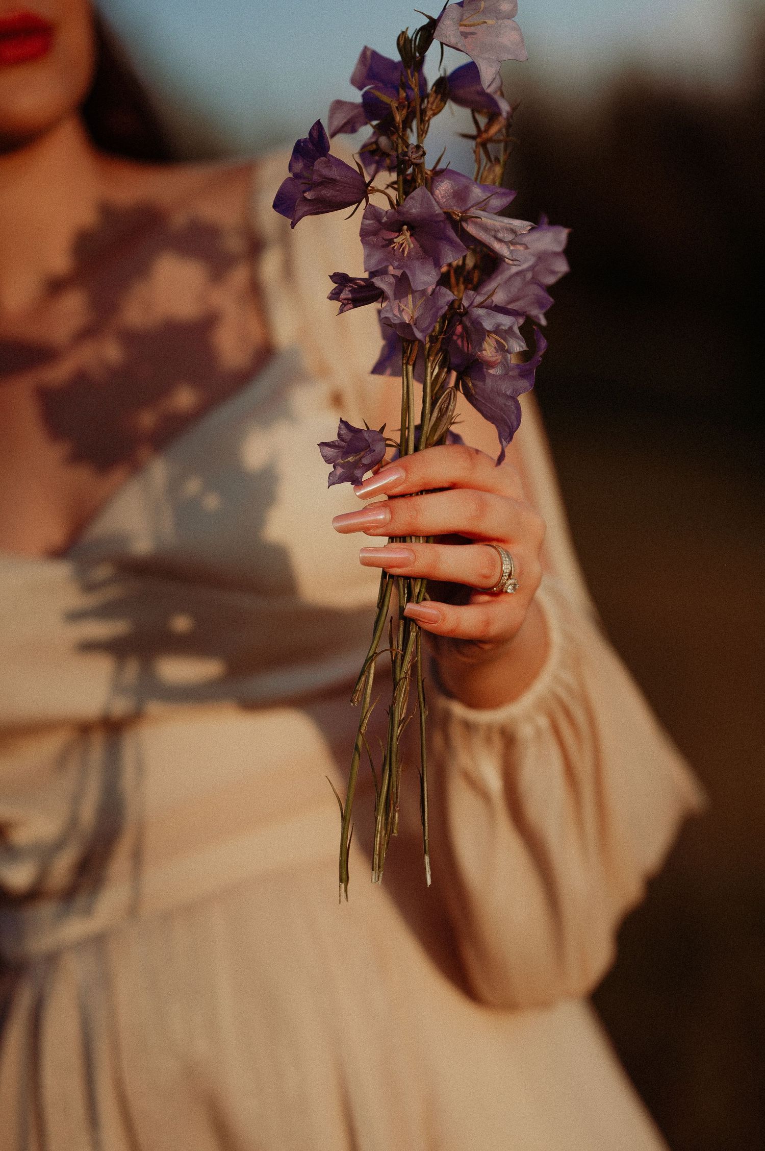 Bride in ivory dress holds delicate purple wildflowers with elegant ring visible in warm sunset light.